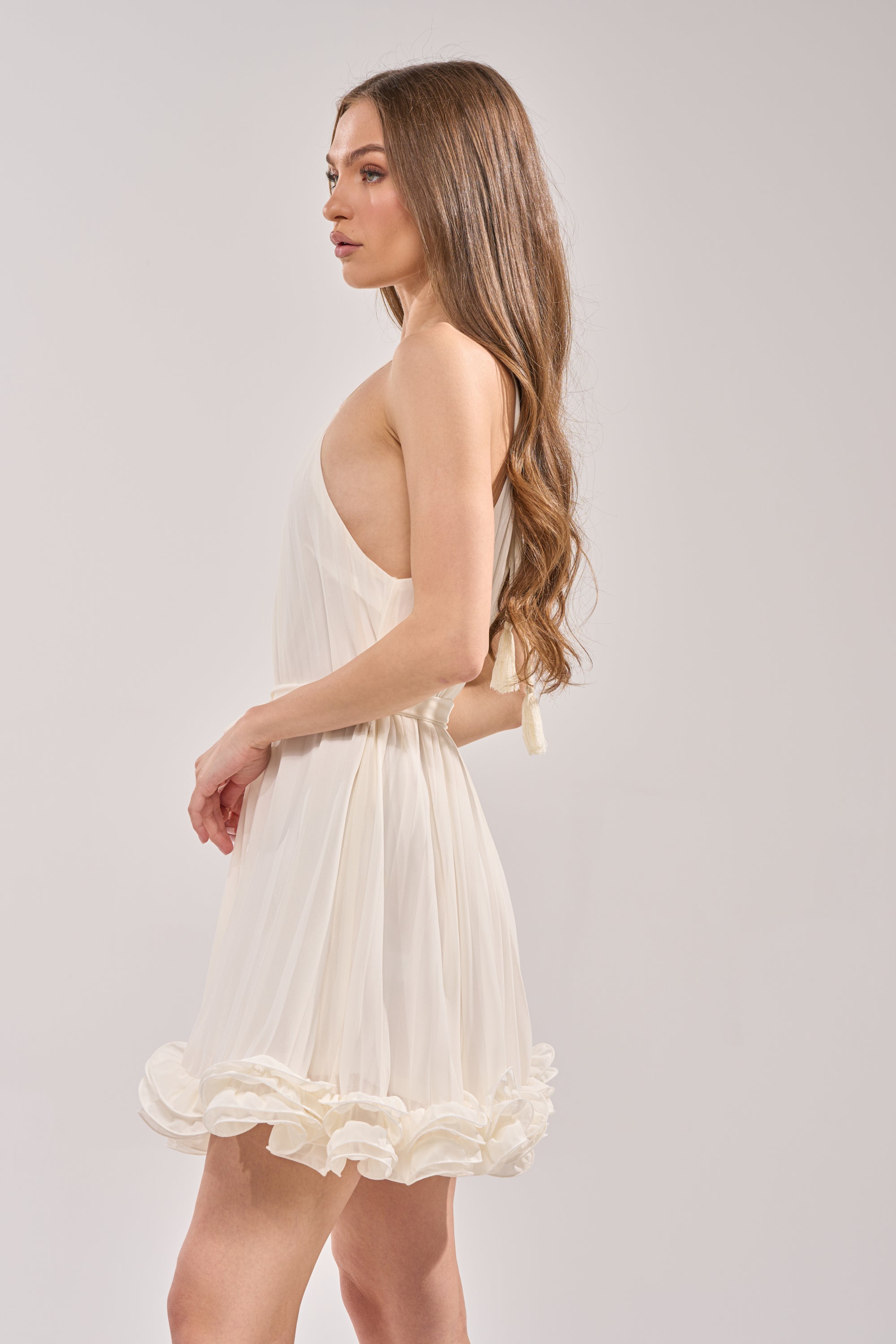 A woman with long, wavy brown hair wears the CAMERA PLEASE RUFFLE MINI DRESS—a sleeveless white mini dress with a ruffled hem and fit-and-flare silhouette—standing sideways against a plain light background. 1