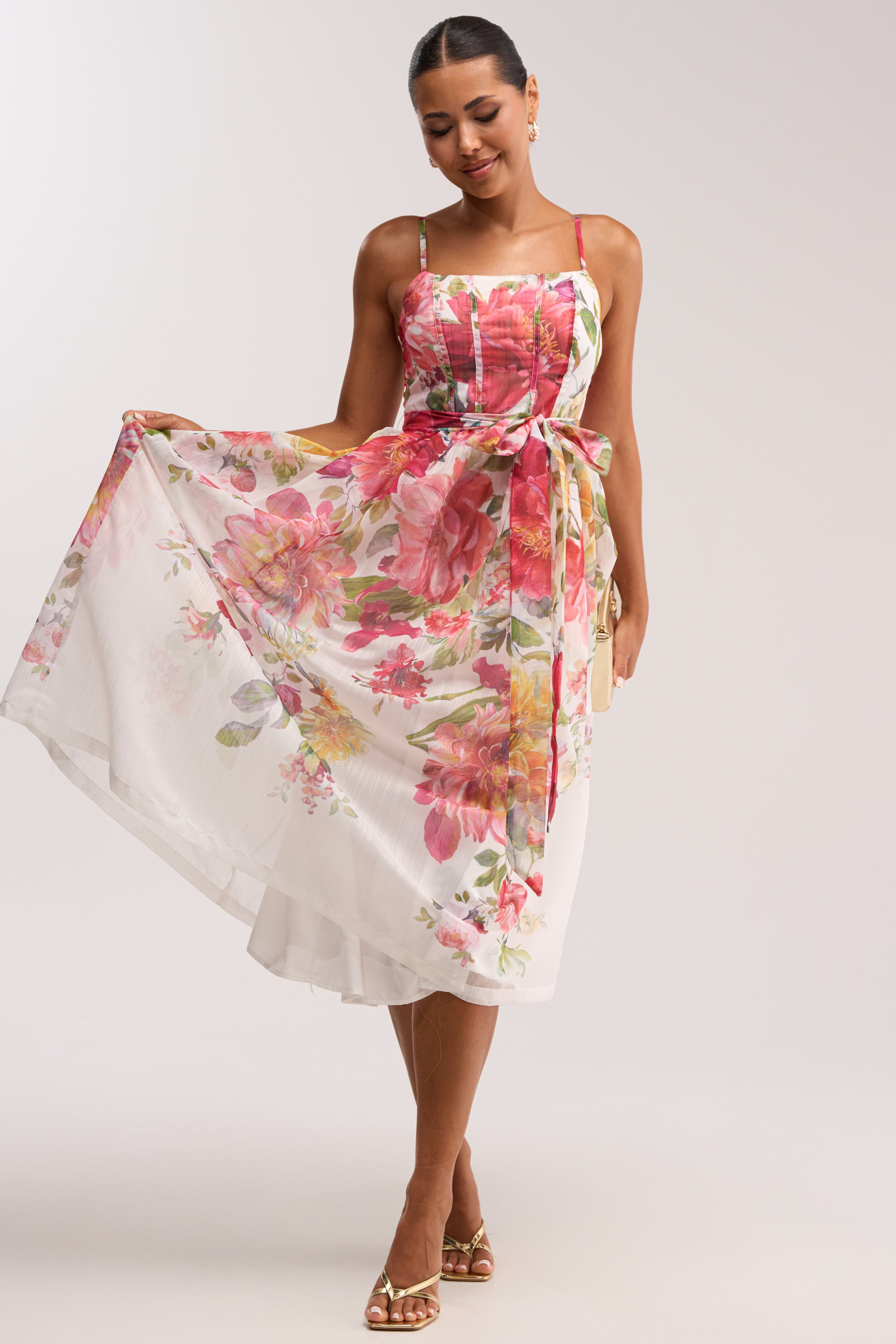 Wearing the GARDEN PARTY FLORAL CHIFFON MIDI DRESS, a sleeveless white midi dress with vibrant pink and orange floral print, she displays the flowing skirt—an elegant option for special occasions. Her hair is tied back and she sports sandals with earrings.