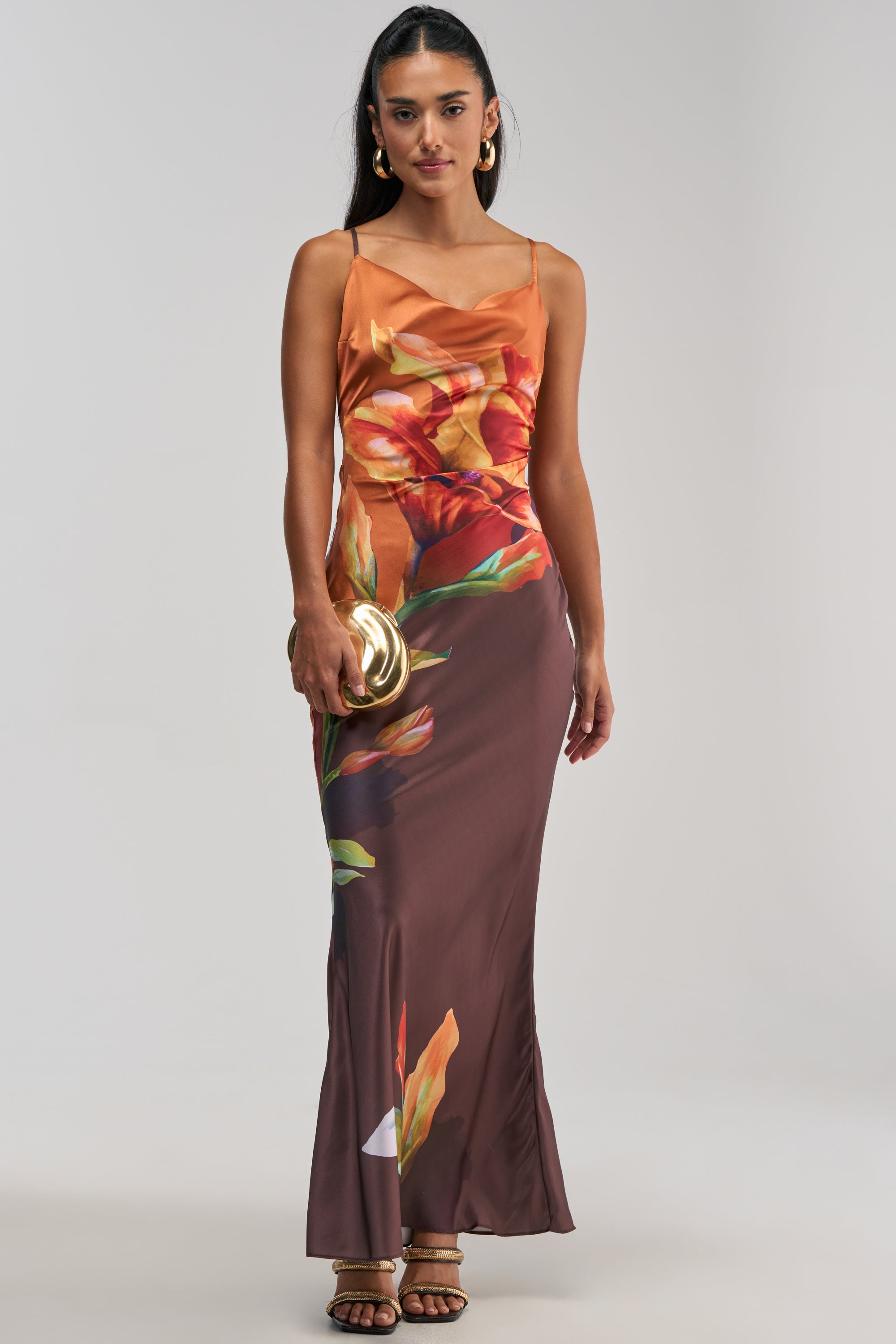 A woman wearing THE MAIN EVENT FLORAL SATIN MAXI DRESS IN ORANGE—a sleeveless, orange satin maxi dress with a brown-toned floral print, ideal for summer date nights or as a wedding guest outfit—holds a gold clutch while standing against a plain light background.