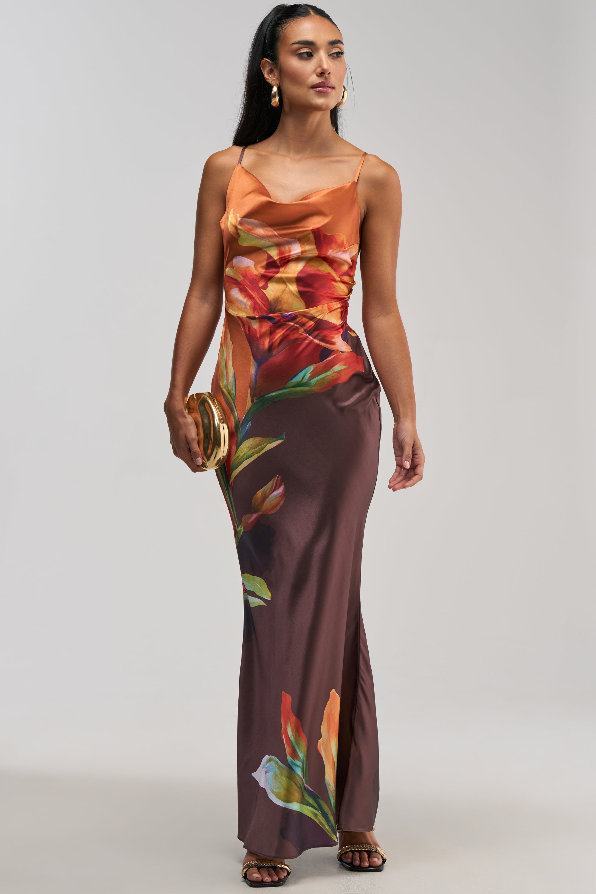 Wearing THE MAIN EVENT FLORAL SATIN MAXI DRESS IN ORANGE—a sleeveless, floor-length satin maxi dress featuring an eye-catching red and orange floral print—she holds a round gold clutch and stands against a plain light background, making this the perfect choice for a summer date night or an elegant wedding guest look.