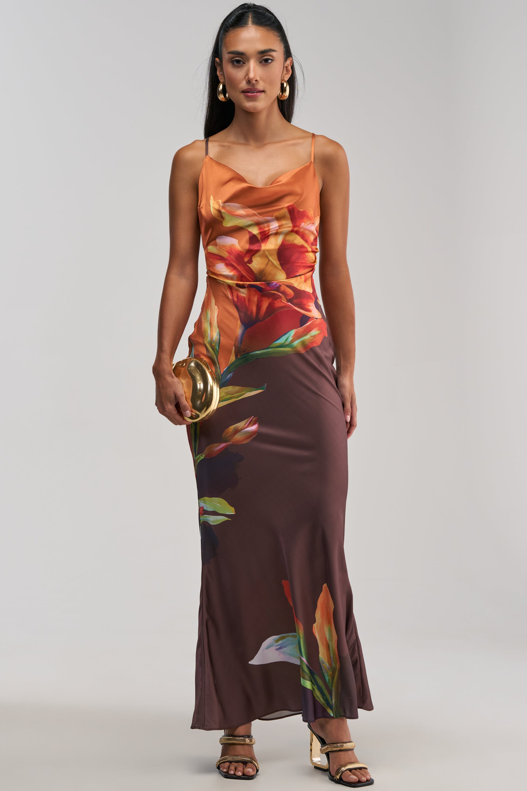 Dressed in THE MAIN EVENT FLORAL SATIN MAXI DRESS IN ORANGE—a stunning orange floral satin maxi with spaghetti straps, featuring an orange bodice and brown skirt—she completes the elegant summer date night or wedding guest look with gold hoop earrings, a gold clutch, and strappy heels.