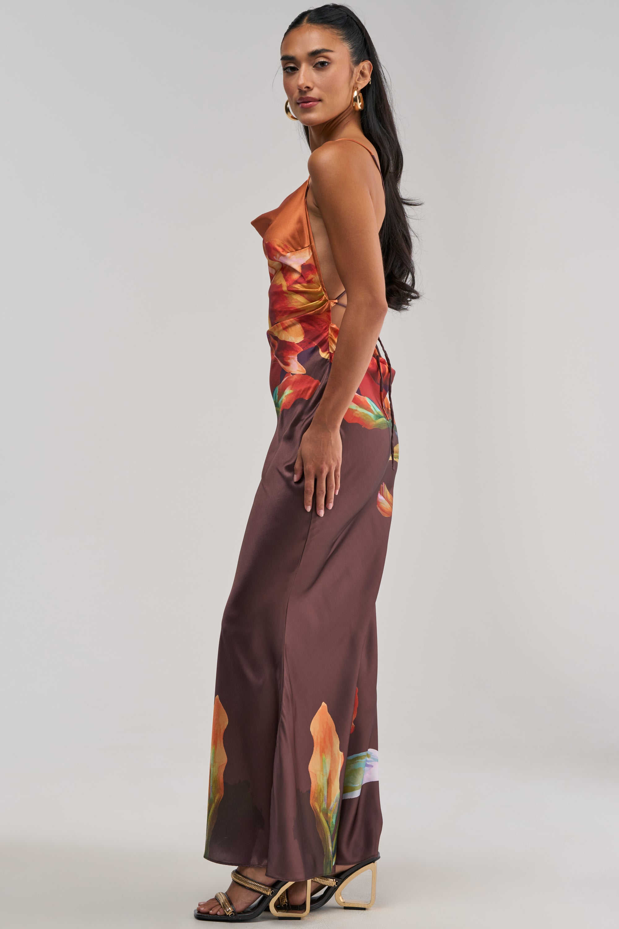 A woman stands in profile against a neutral background, wearing THE MAIN EVENT FLORAL SATIN MAXI DRESS IN ORANGE—a sleeveless, floor-length silhouette featuring a vibrant floral print in shades of orange and brown. Styled with gold earrings and heeled sandals, it’s an elegant choice for a summer date night or as a wedding guest dress.