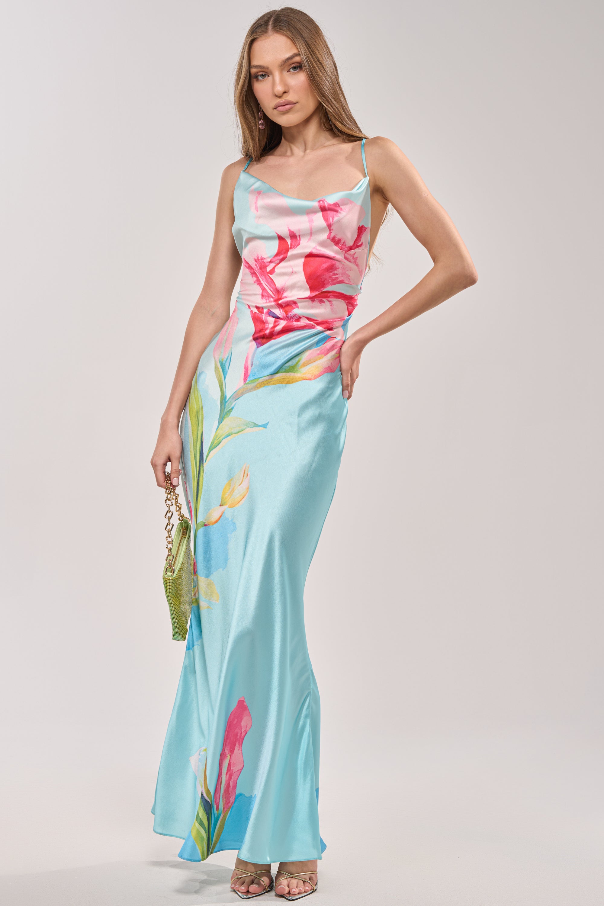 Wearing THE MAIN EVENT FLORAL SATIN MAXI DRESS—featuring colorful floral prints, a fitted waist, and spaghetti straps—a woman poses with a green handbag and heeled sandals, perfect for summer weddings. 3