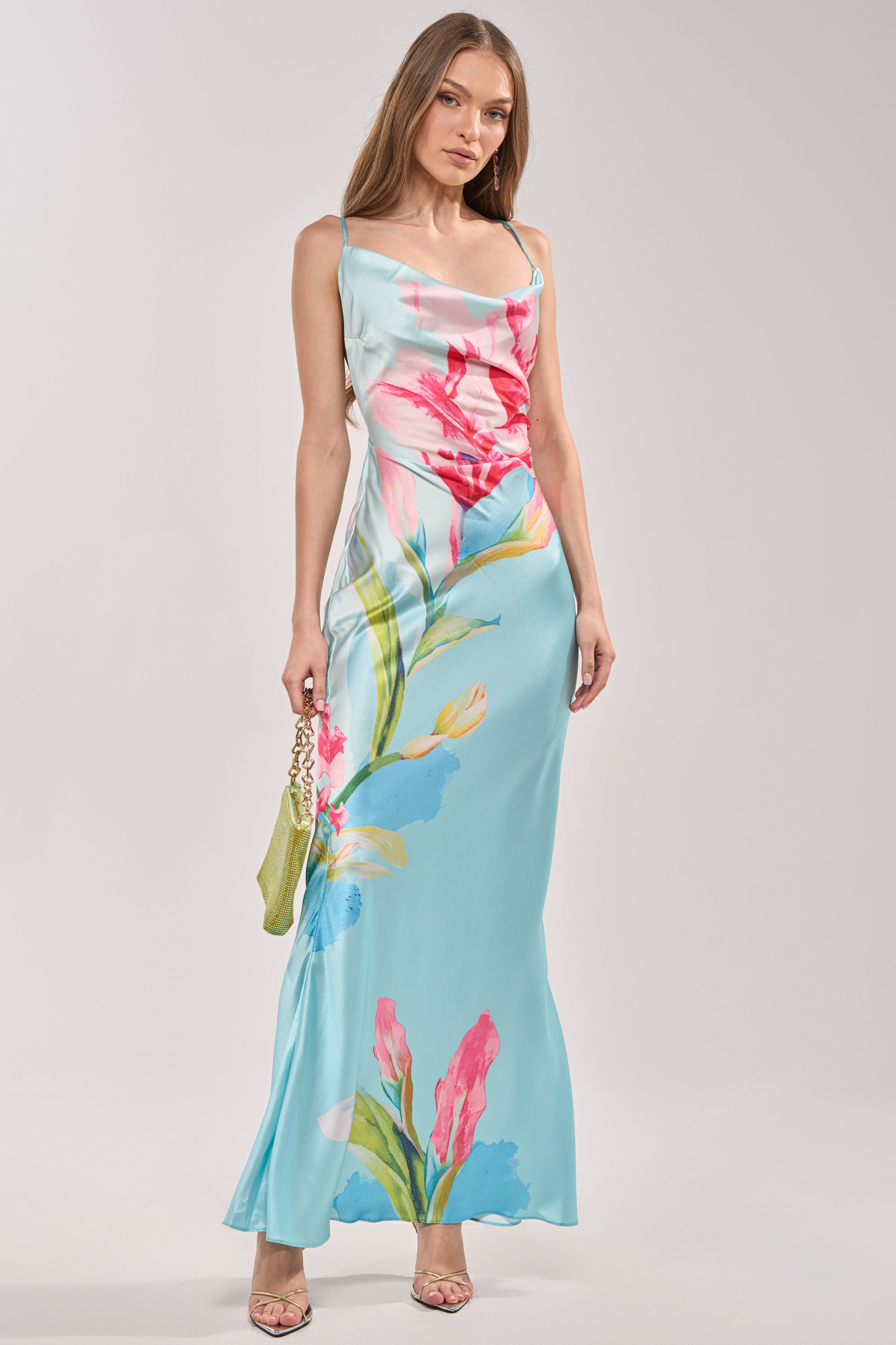 Model wears THE MAIN EVENT FLORAL SATIN MAXI DRESS in light blue with colorful floral prints, styled with a green handbag and open-toe heels—an ideal tropical vacation look against a plain background. 4