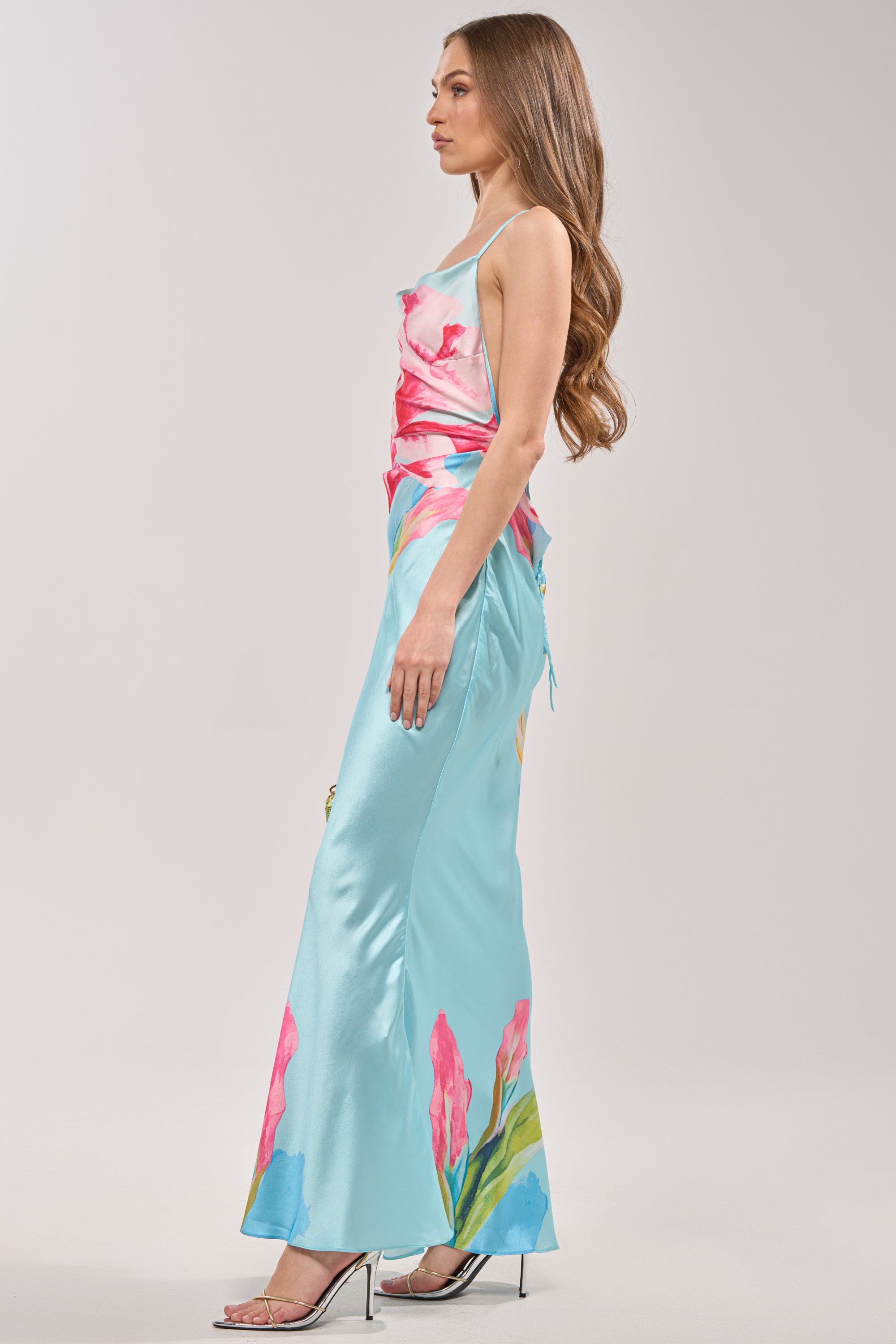 Wearing THE MAIN EVENT FLORAL SATIN MAXI DRESS, a woman with long wavy hair and silver heels stands in profile against a light background—the perfect tropical vacation look. 5