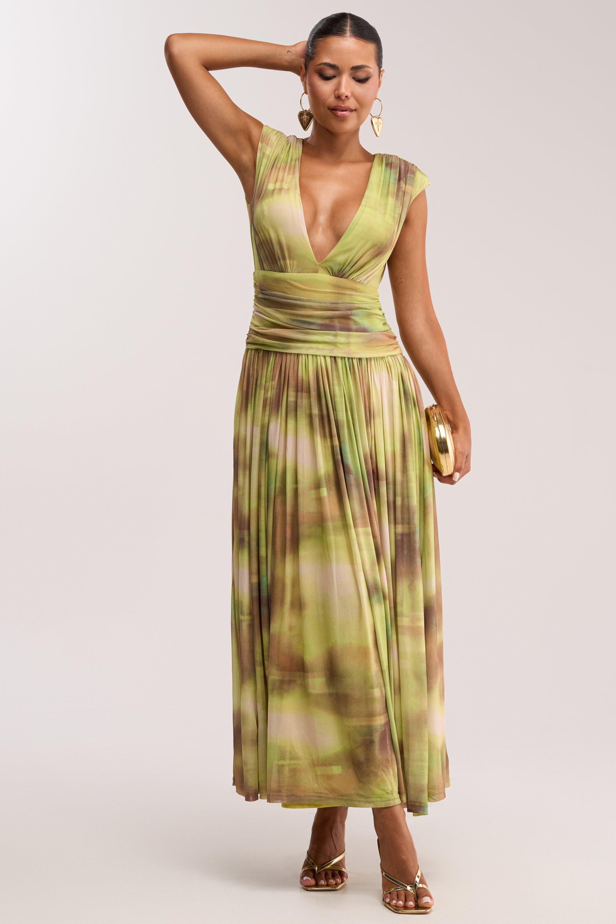 A woman poses against a plain light background in the TEA TIME ABSTRACT MAXI DRESS IN GREEN with cap sleeves, deep V-neckline, gold sandals, heart-shaped earrings, and a small gold clutch—a perfect summer date night look.