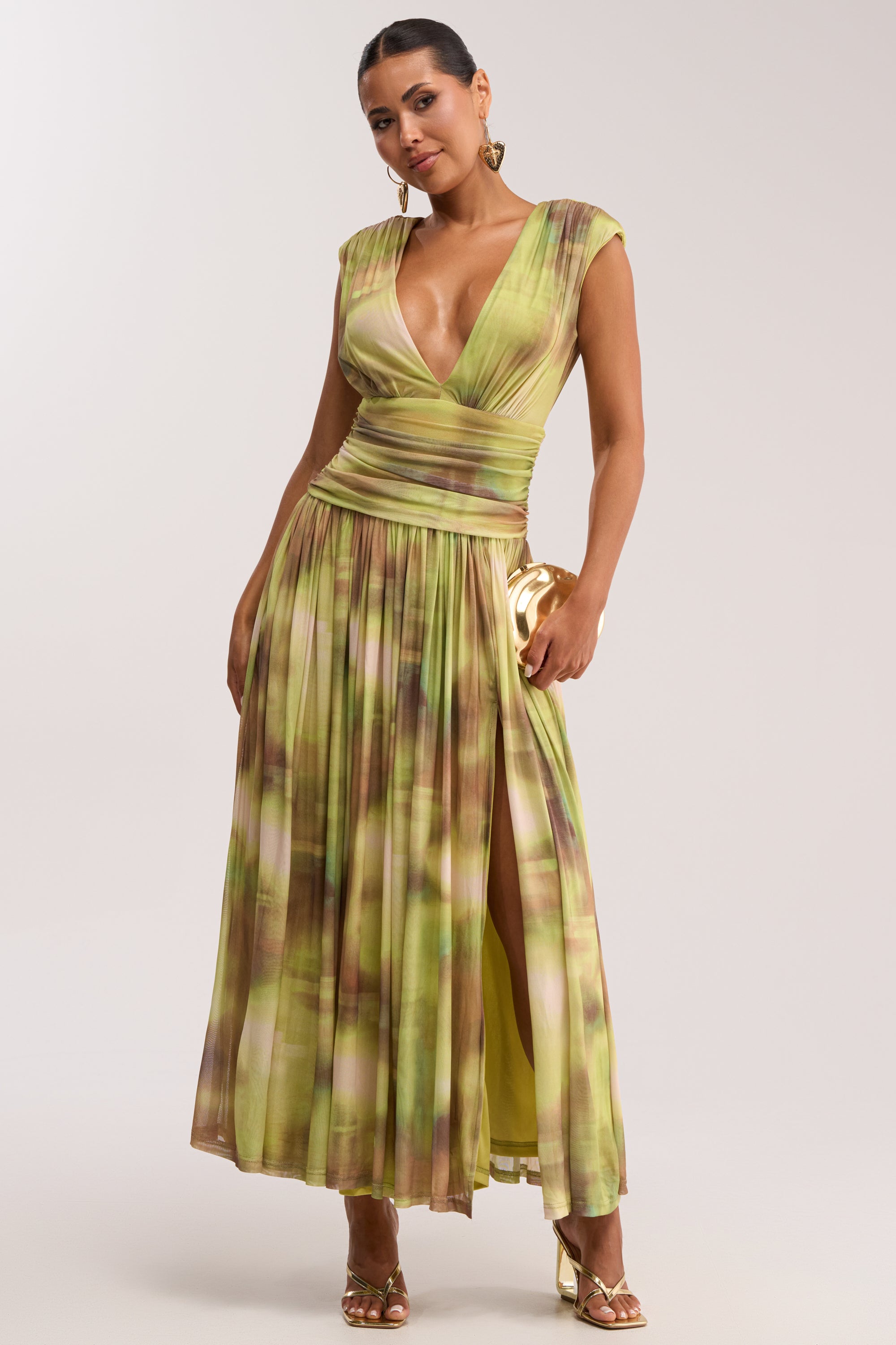 A woman poses against a neutral background in the TEA TIME ABSTRACT MAXI DRESS IN GREEN, an elegant plunge maxi dress with a green abstract print, deep V-neck, cap sleeves, gold earrings, metallic heels, and a gold clutch.