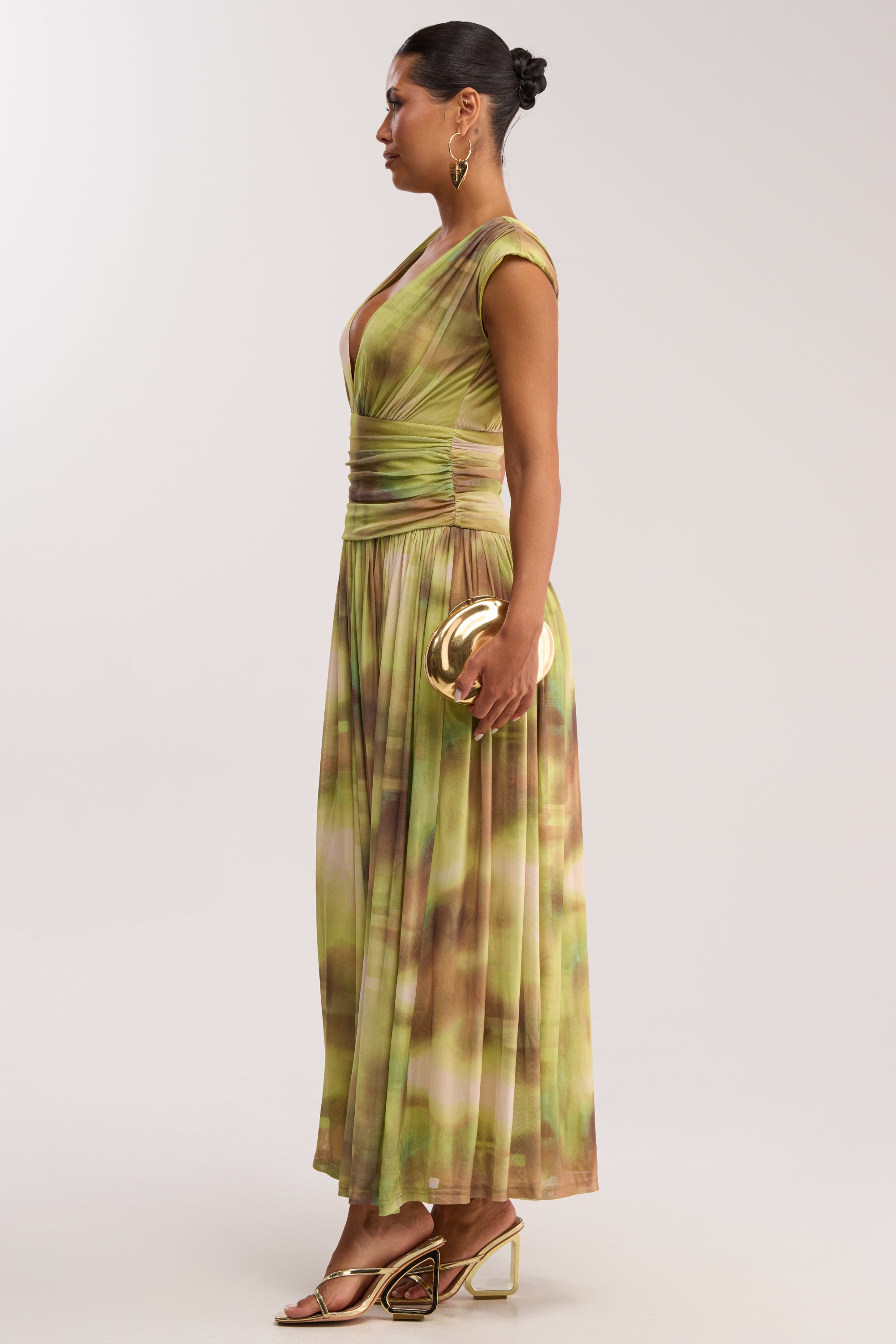 A woman in profile wears the TEA TIME ABSTRACT MAXI DRESS IN GREEN, featuring a deep V-neck, ruched waist, and cap sleeves. She accessorizes with a metallic gold clutch, gold heels, hoop earrings, and a neat bun.