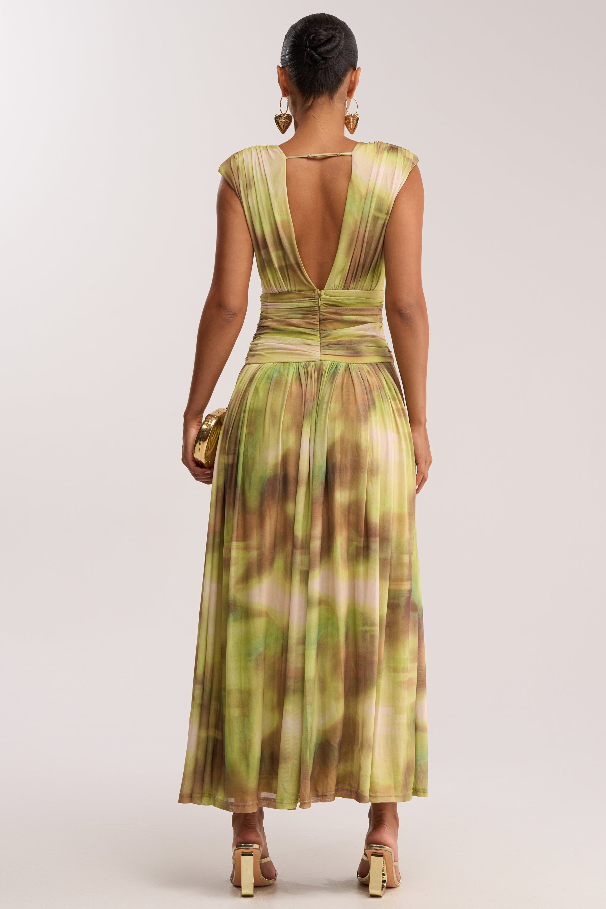 Woman stands with her back to the camera, wearing the TEA TIME ABSTRACT MAXI DRESS IN GREEN featuring pleats, a deep V-back, and cap sleeves—styled with gold heels, hoop earrings, and a gold clutch for a chic summer date night look.