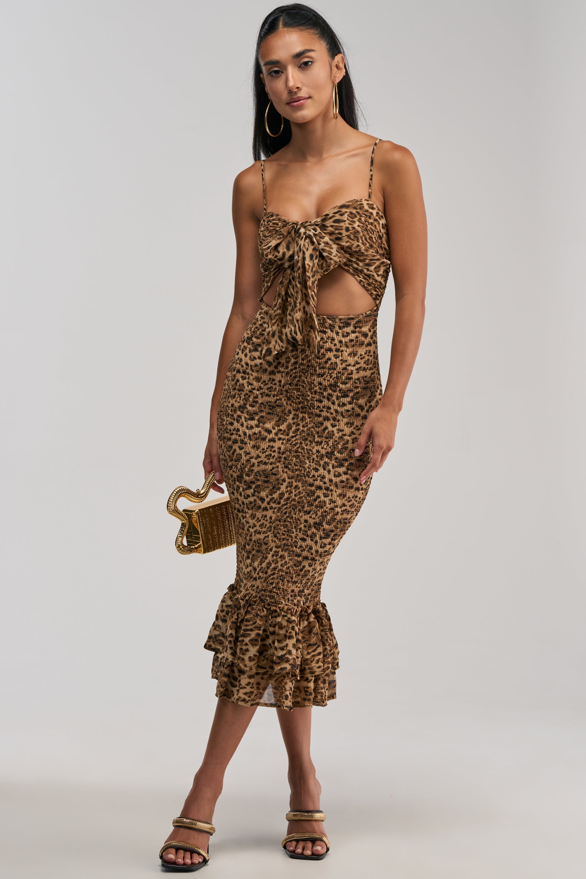 Styled in the BODY-ODY LEOPARD MIDI DRESS, this look features a leopard print chiffon fabric with a front tie, waist cutout detail, and ruffled hem. The chic midi dress is paired with a small gold clutch, hoop earrings, and strappy heels.