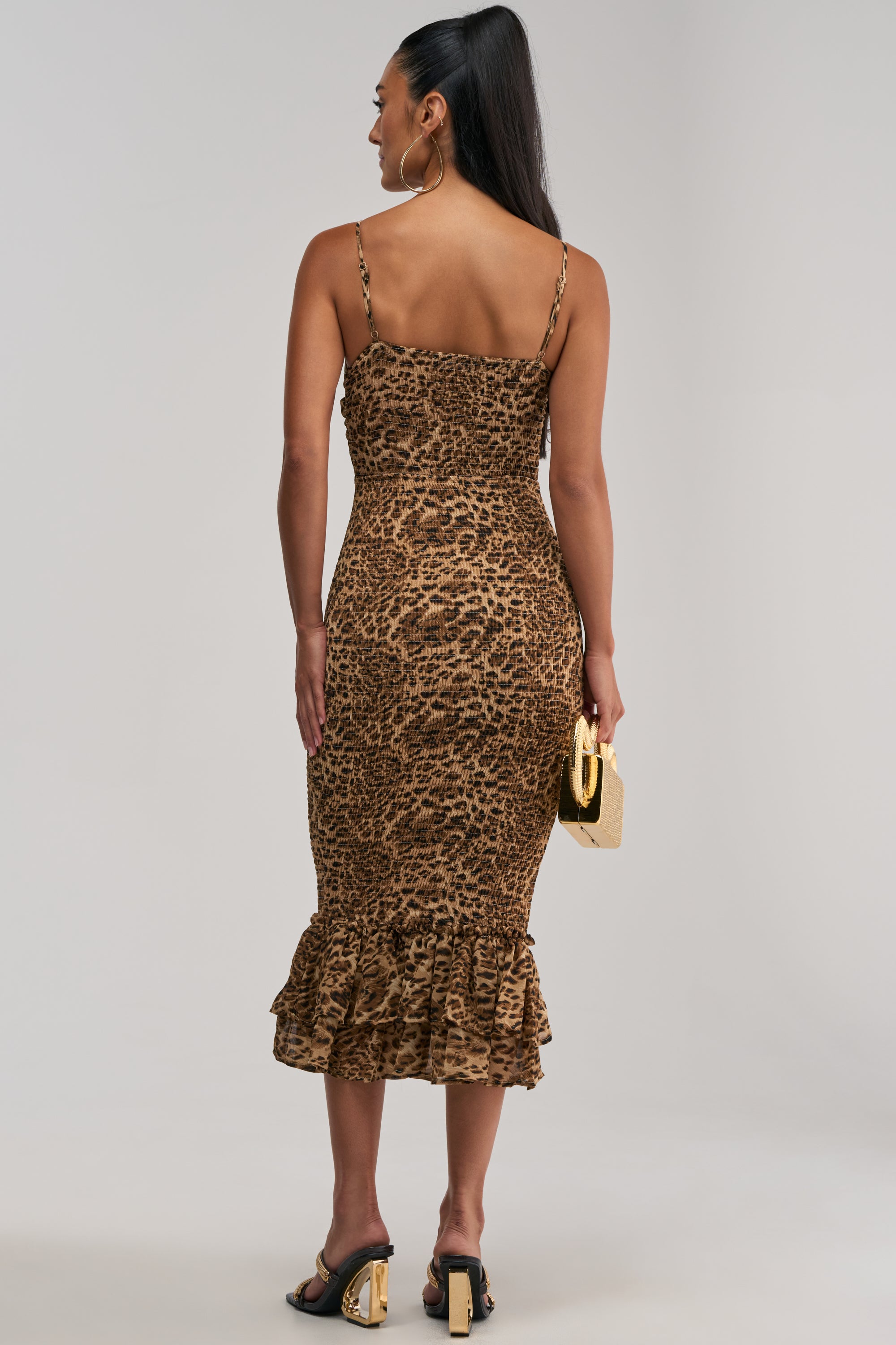 A woman faces away, wearing the BODY-ODY LEOPARD MIDI DRESS—a fitted chiffon midi dress featuring a leopard print, ruffled hem, and thin straps. She accessorizes with hoop earrings, a small gold clutch, and black heeled sandals, with her hair styled in a high ponytail.