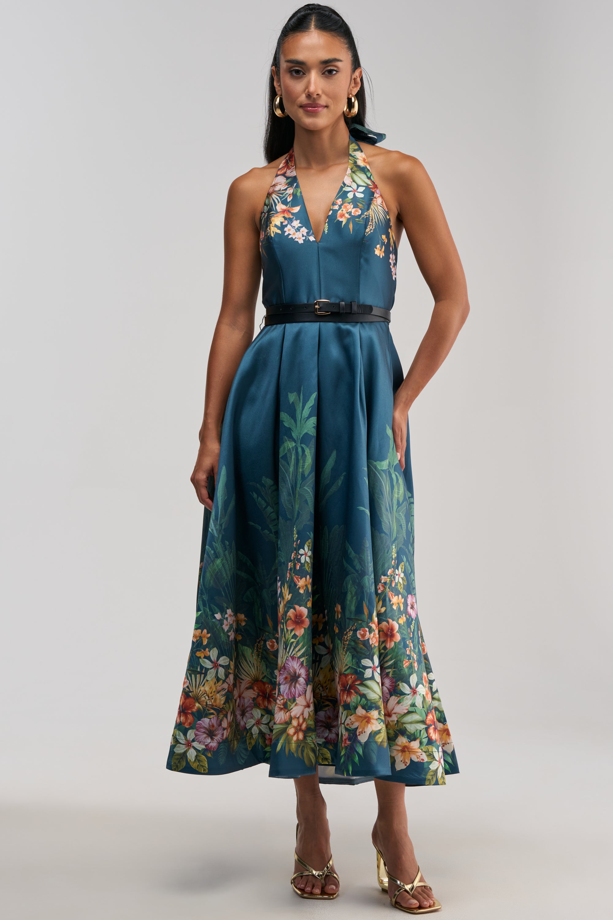 A woman stands against a plain light background, styled for a chic summer date night in the BOOKED AND BUSY FLORAL MIDI DRESS IN BLUE MULTI—a sleeveless, floral print halter dress with vibrant teal-blue hues—paired with a black belt, gold hoop earrings, and gold open-toe heels.