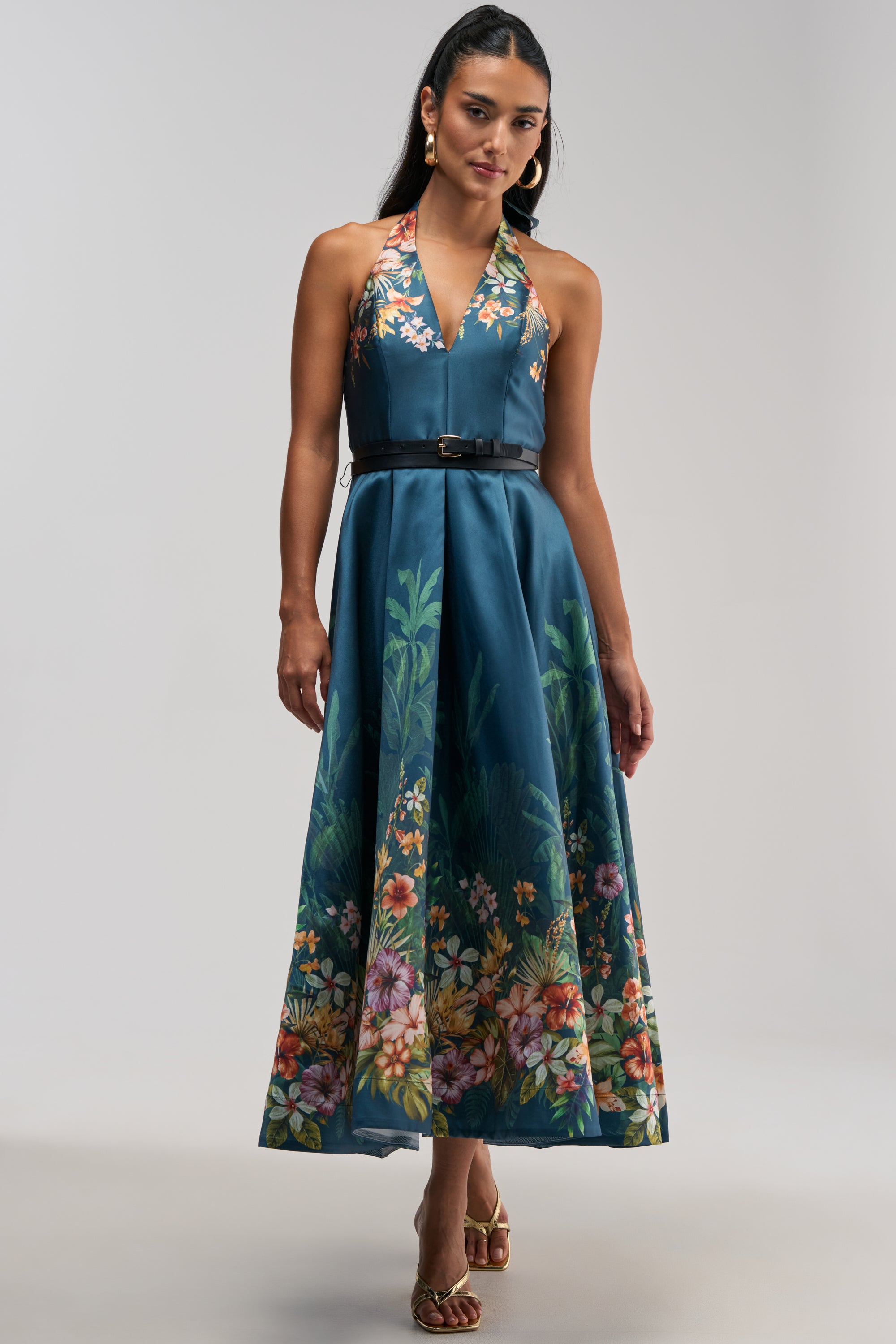 A woman wears the BOOKED AND BUSY FLORAL MIDI DRESS IN BLUE MULTI, a sleeveless satin halter dress with a floral pattern along the hem, paired with a black belt at the waist, hoop earrings, and gold sandals, standing against a plain light background.