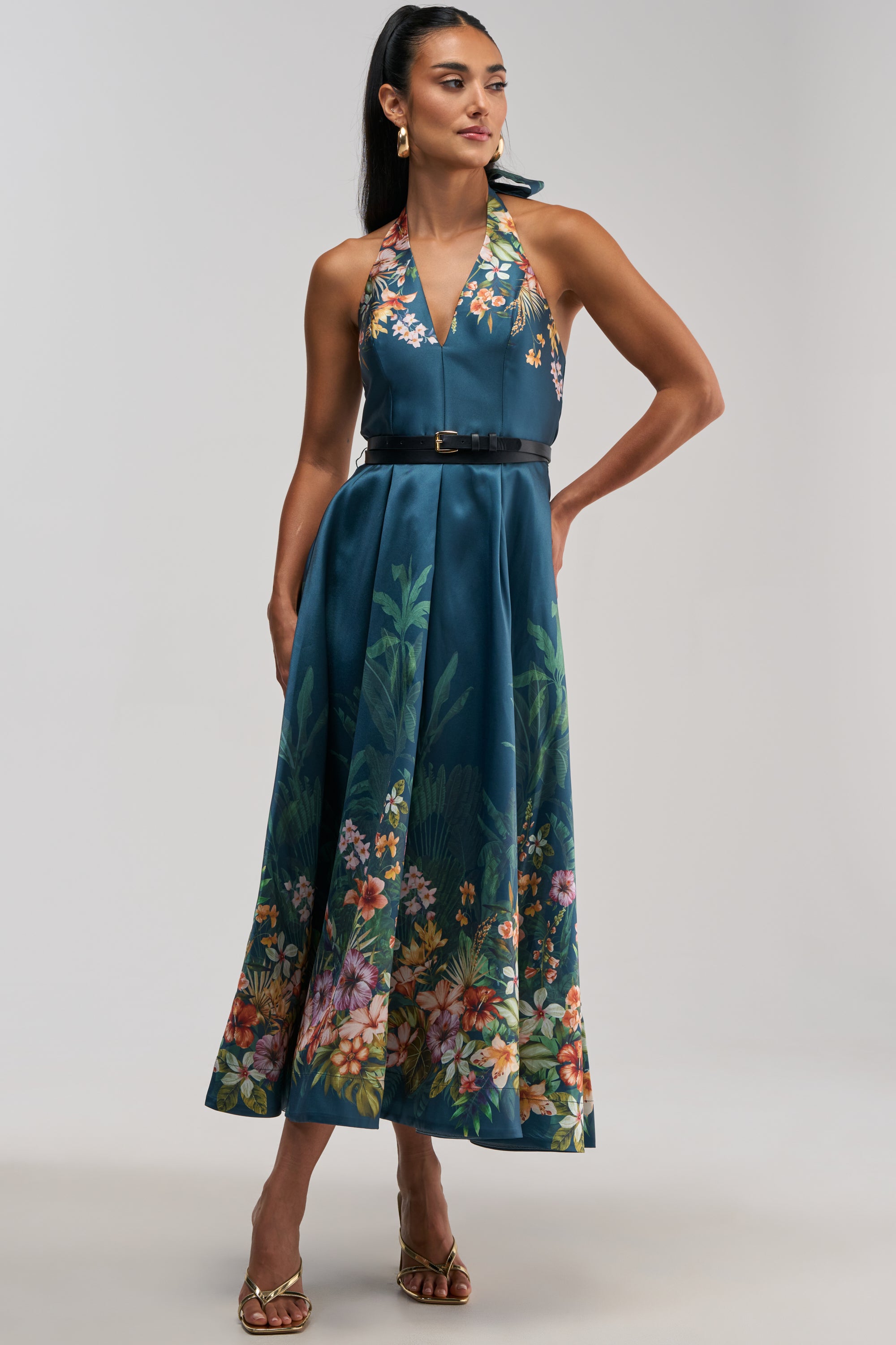 Styled in the BOOKED AND BUSY FLORAL MIDI DRESS IN BLUE MULTI—a sleeveless, halter-neck dress featuring a beautiful floral pattern along the hem—this look is perfect for a summer date night. The outfit is completed with a black belt, gold heels, and hoop earrings, set against a plain light background.