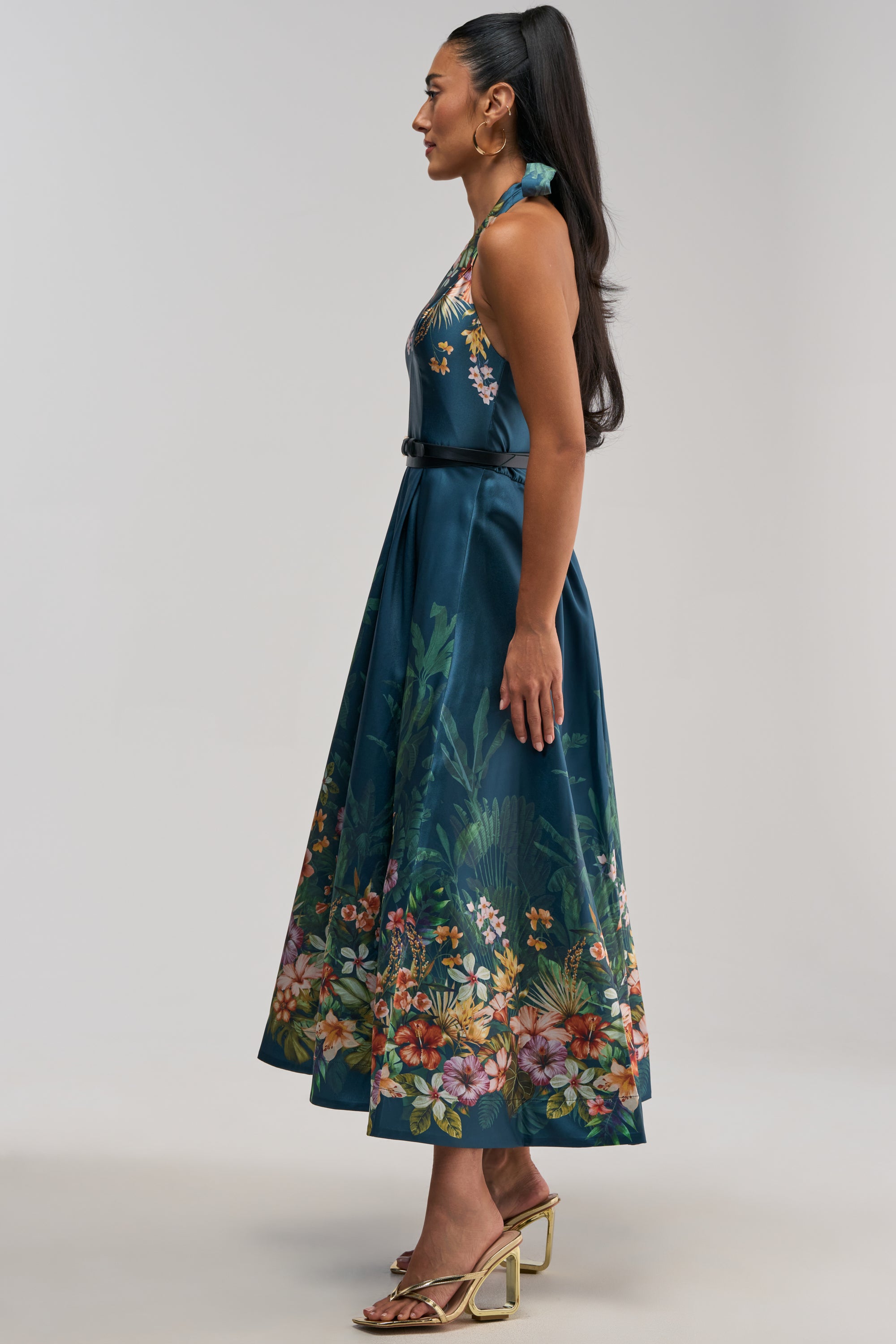 A woman wearing the BOOKED AND BUSY FLORAL MIDI DRESS IN BLUE MULTI—a sleeveless floral dress with a flared skirt—stands in profile on gold heeled sandals against a plain light background.