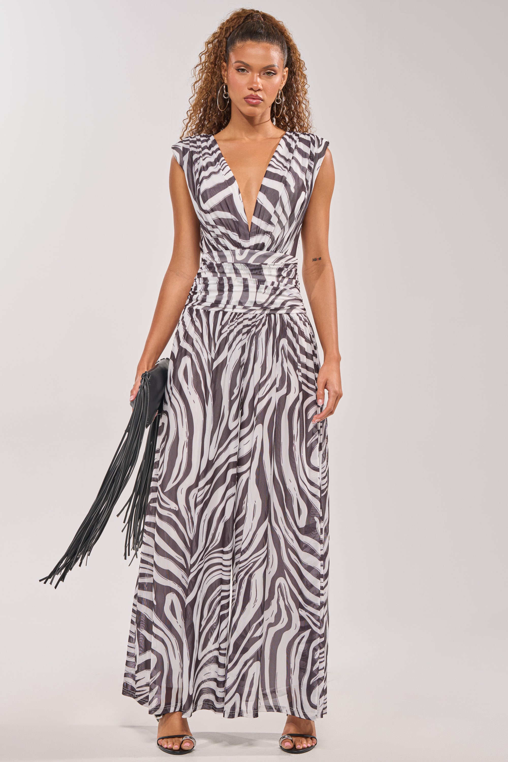 A woman wears the Jungle Fever Zebra Print Jumpsuit, a sleeveless v-neck, floor-length piece with a gathered waist. She holds a black fringed clutch and stands in high-heeled sandals against a plain background. 0