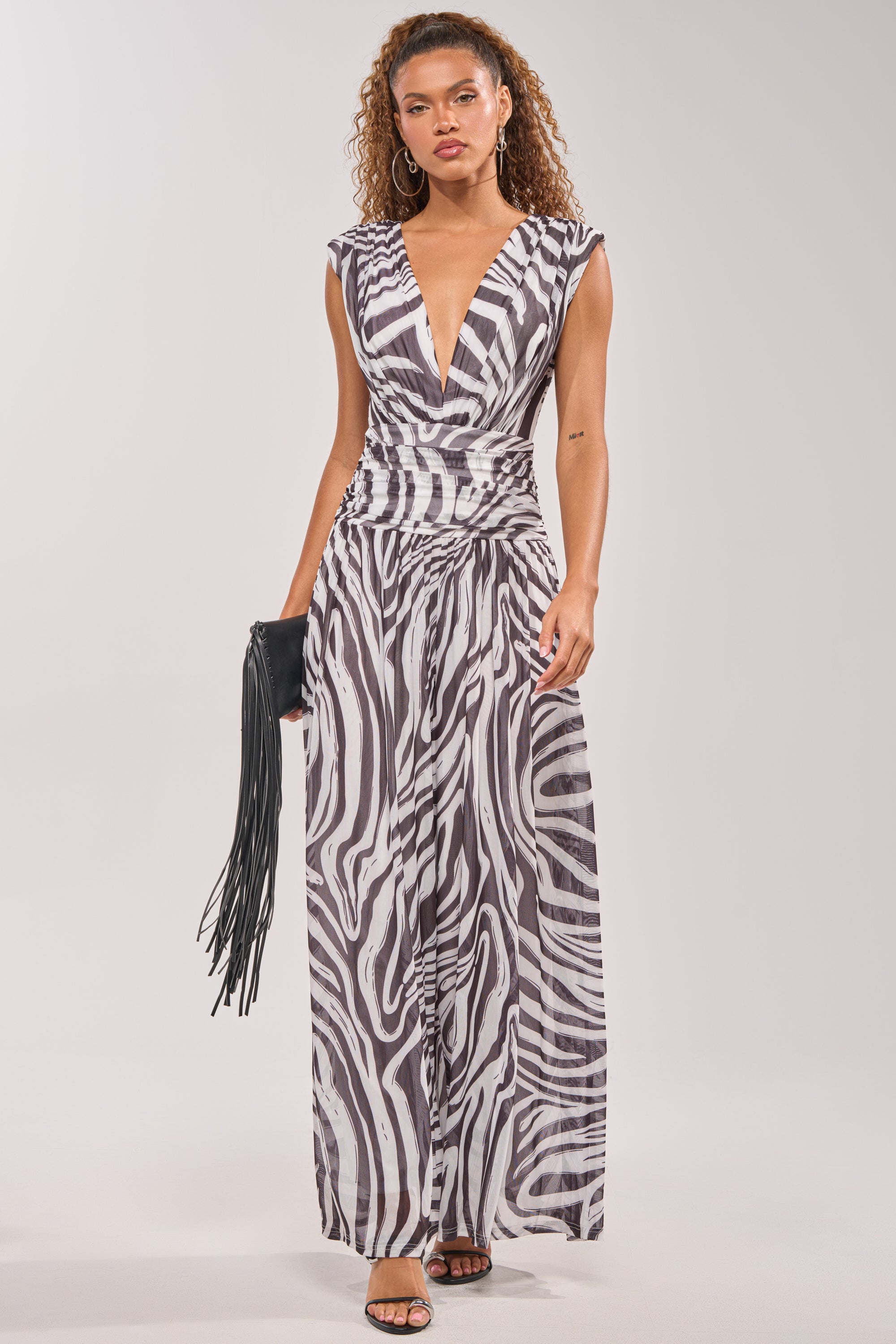 A woman wears the JUNGLE FEVER ZEBRA PRINT JUMPSUIT—sleeveless with a deep V-neck—paired with hoop earrings, black heels, and a fringed black clutch. 1