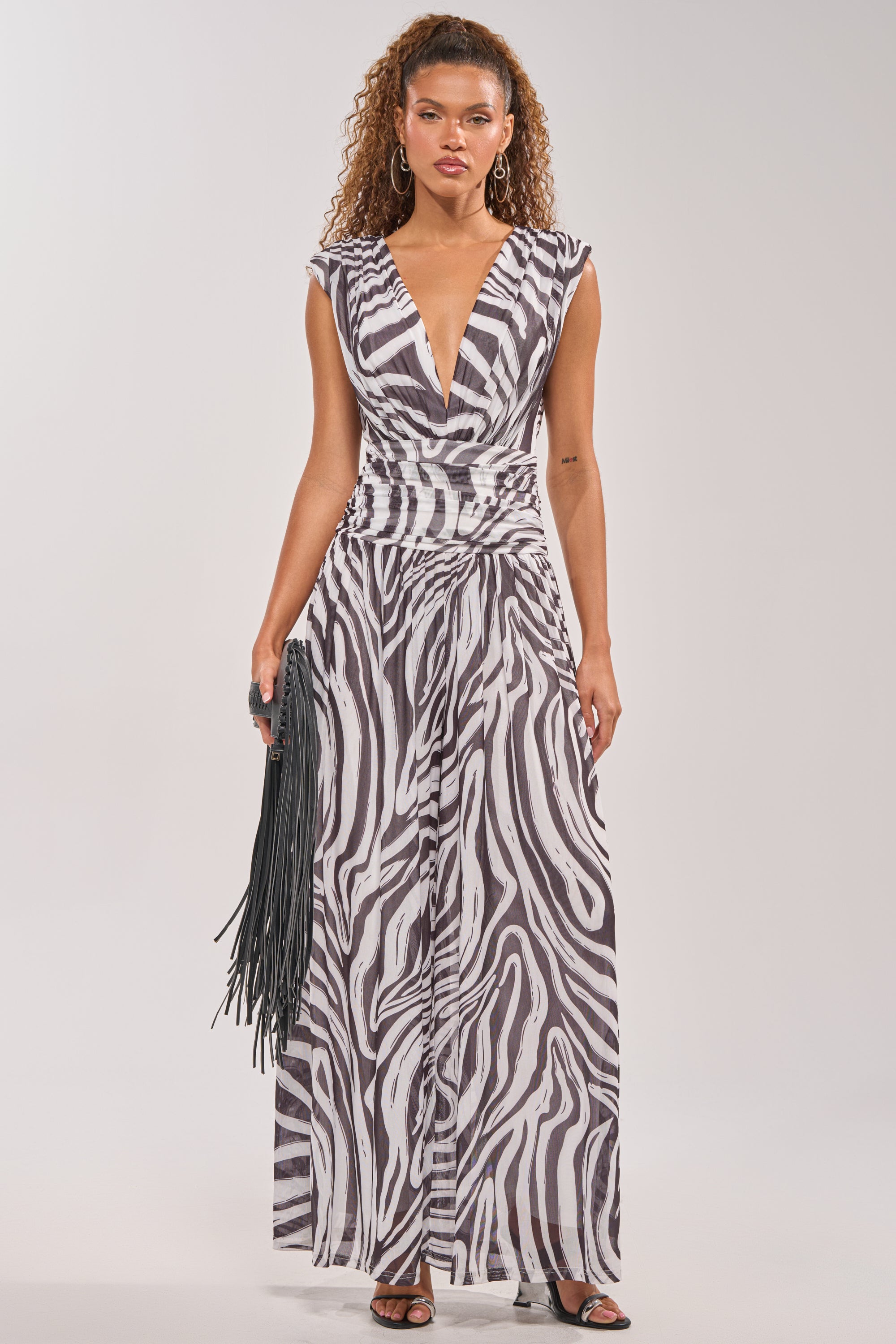 A woman with curly hair models the JUNGLE FEVER ZEBRA PRINT JUMPSUIT, featuring a deep V-neck and wide legs, styled with a black fringed bag and sandals, against a plain light background. 2