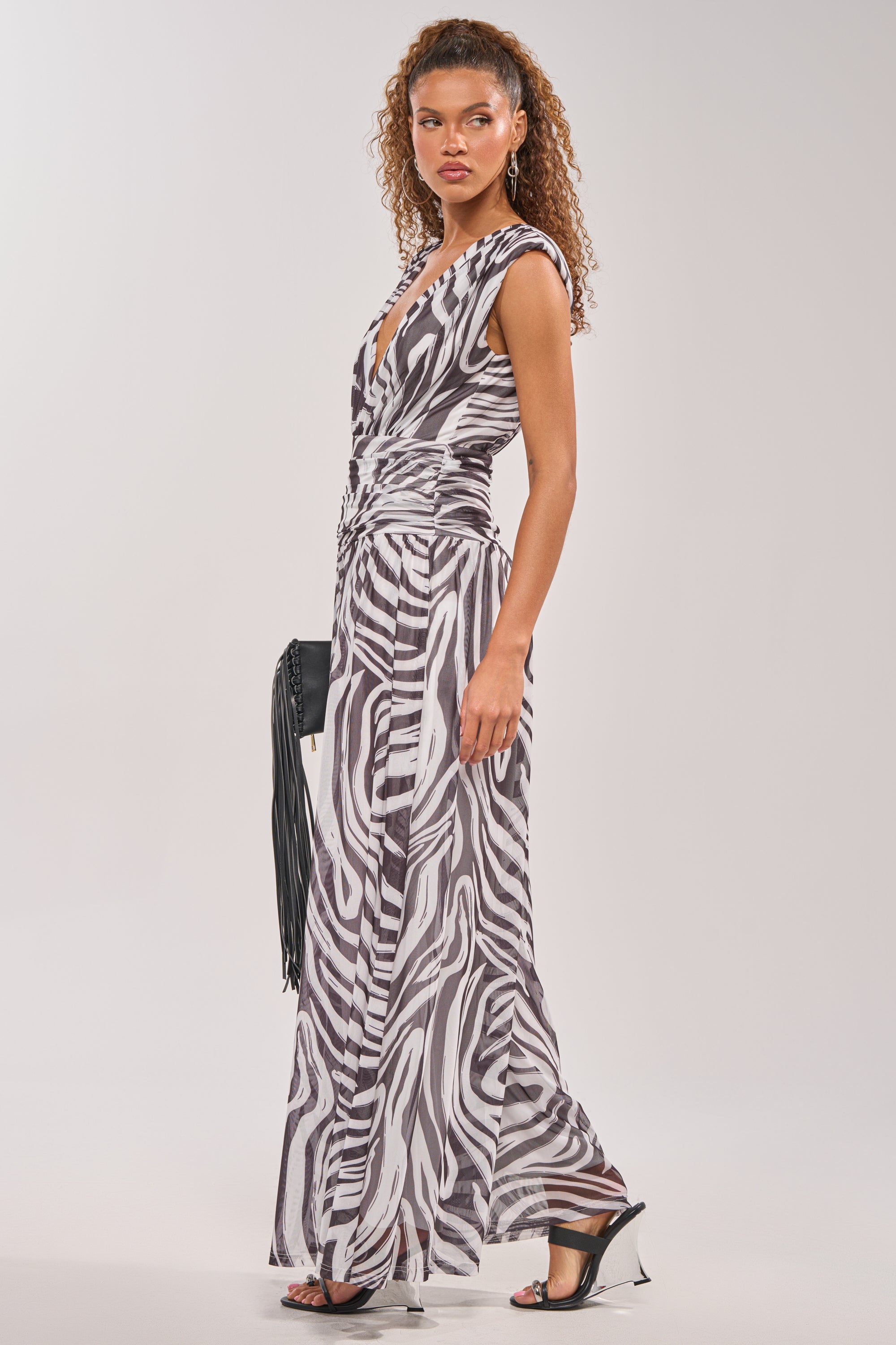 A woman poses against a plain background in the JUNGLE FEVER ZEBRA PRINT JUMPSUIT, a sleeveless, floor-length piece with a deep V-neck, styled with black strappy heels and a black fringed clutch. 3