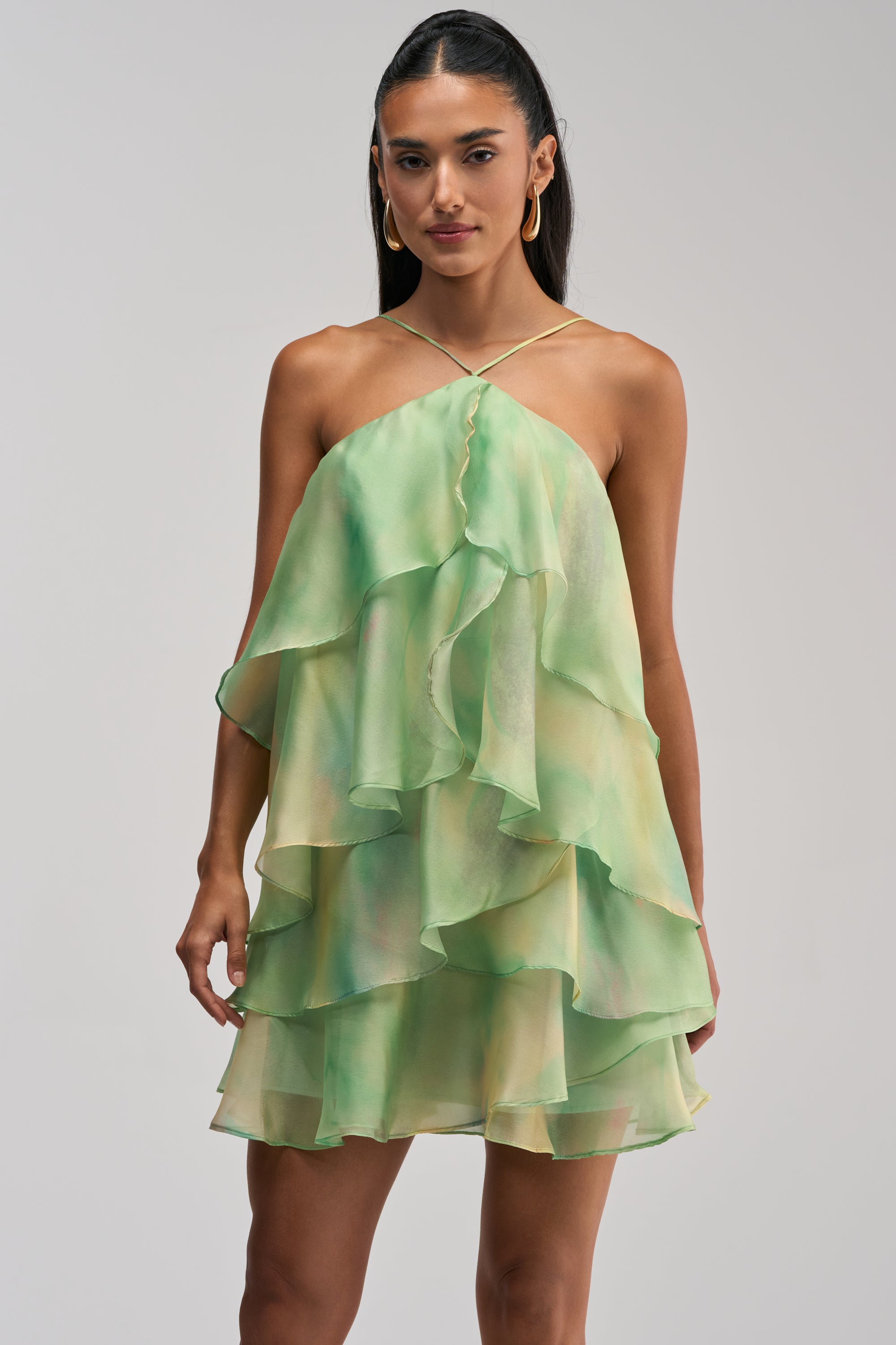 A model wears the Shine Brighter Baby Mini Dress in Green, a green and yellow ombre chiffon mini dress featuring tiered ruffles and thin straps. She pairs it with gold hoop earrings and styles her dark hair pulled back, creating a stunning look that’s perfect for a wedding guest.