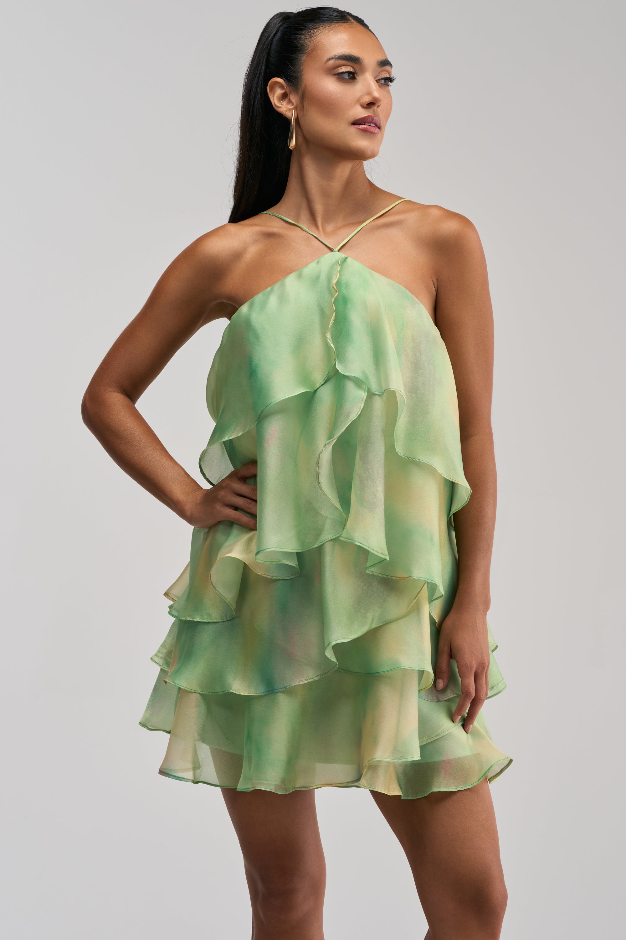 A woman stands with one hand on her hip against a plain light background, wearing the SHINE BRIGHTER BABY MINI DRESS IN GREEN—a sleeveless, layered chiffon mini dress featuring green abstract prints and ruffled tiers, making it an elegant choice for a wedding guest.