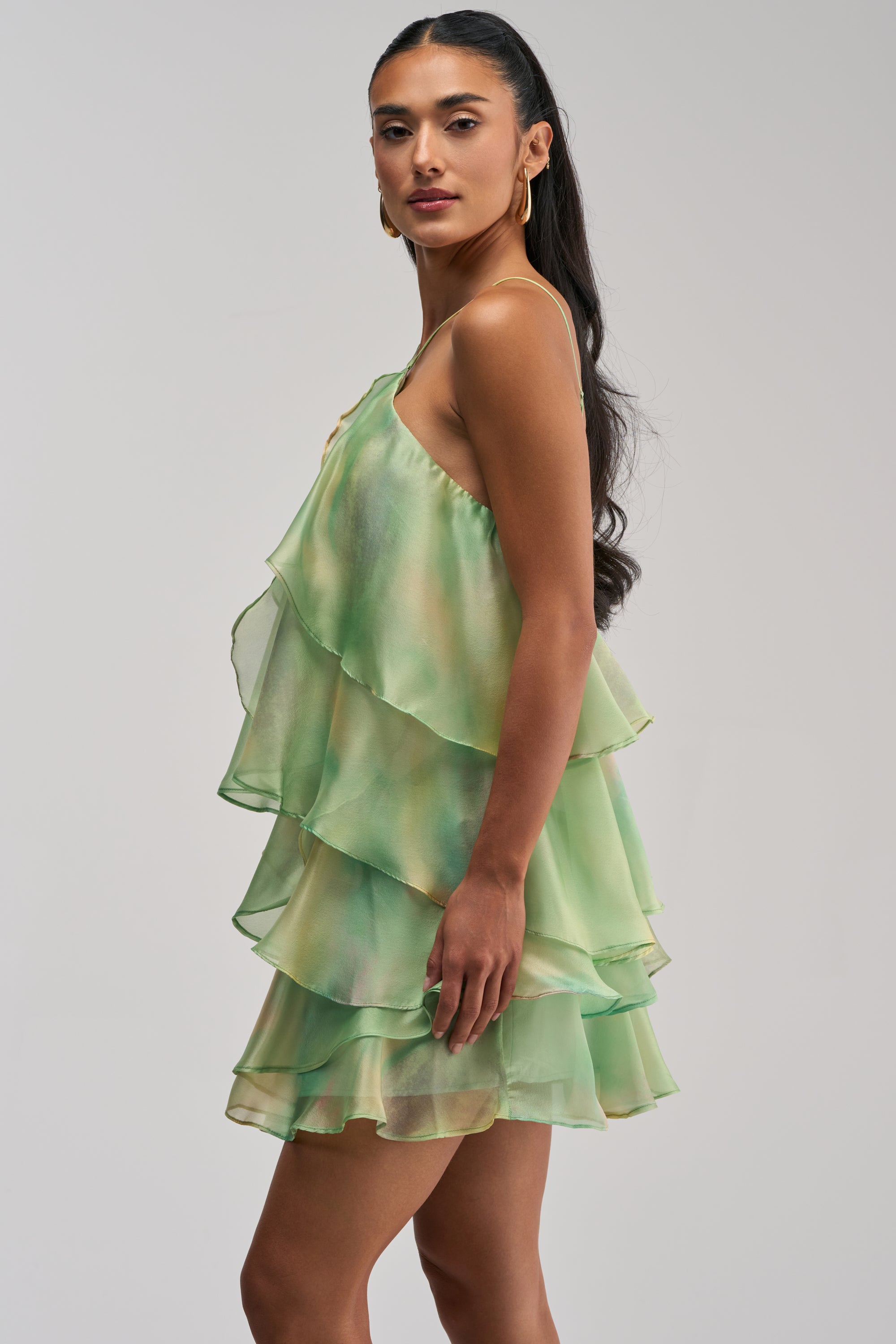 A woman in the SHINE BRIGHTER BABY MINI DRESS IN GREEN—a light green, tiered ruffle mini dress featuring a one-shoulder design—stands sideways and looks at the camera. She has long, wavy hair and accessorizes with gold hoop earrings, making this the perfect wedding guest dress for any special occasion.