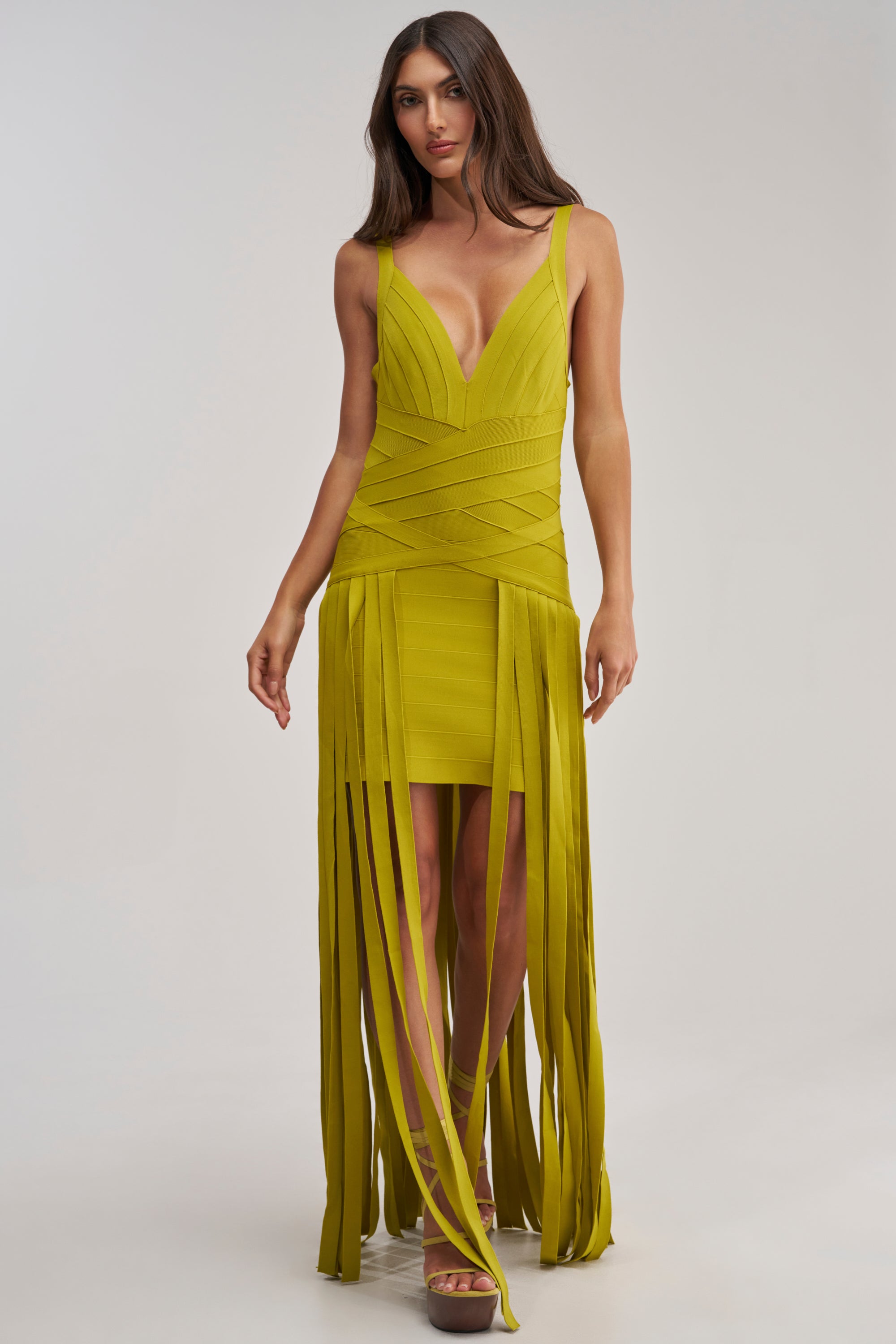 A woman stands against a plain light background wearing the LUCKY CHARM FRINGE MINI DRESS—a chartreuse bandage mini dress featuring a deep V-neckline and long, fringe-like strips on the skirt.