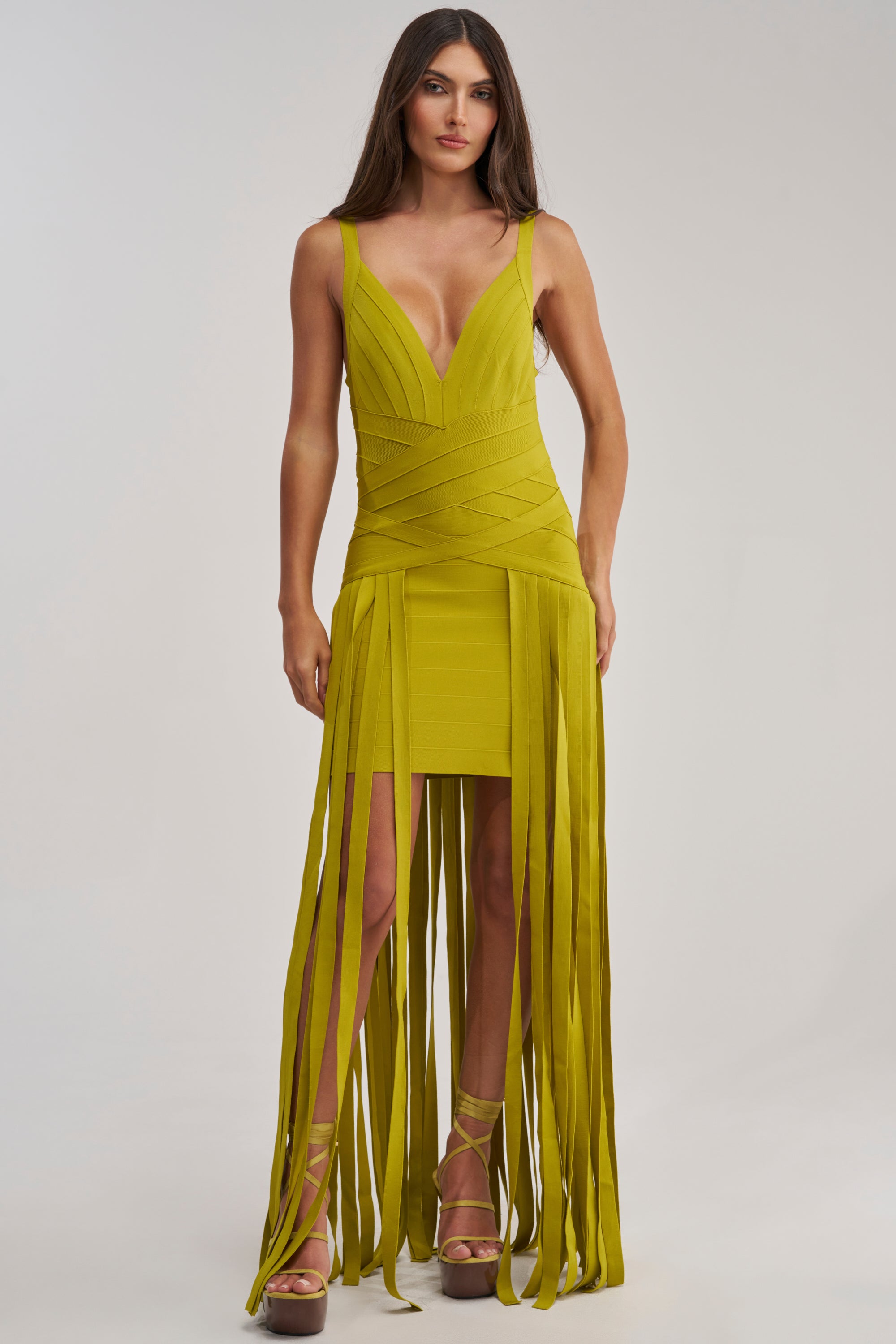 A woman poses against a plain background in the LUCKY CHARM FRINGE MINI DRESS—a sleeveless, chartreuse bandage mini dress with a deep V-neck, long vertical fringe accents, and gold platform heels—ideal for summer soirees.