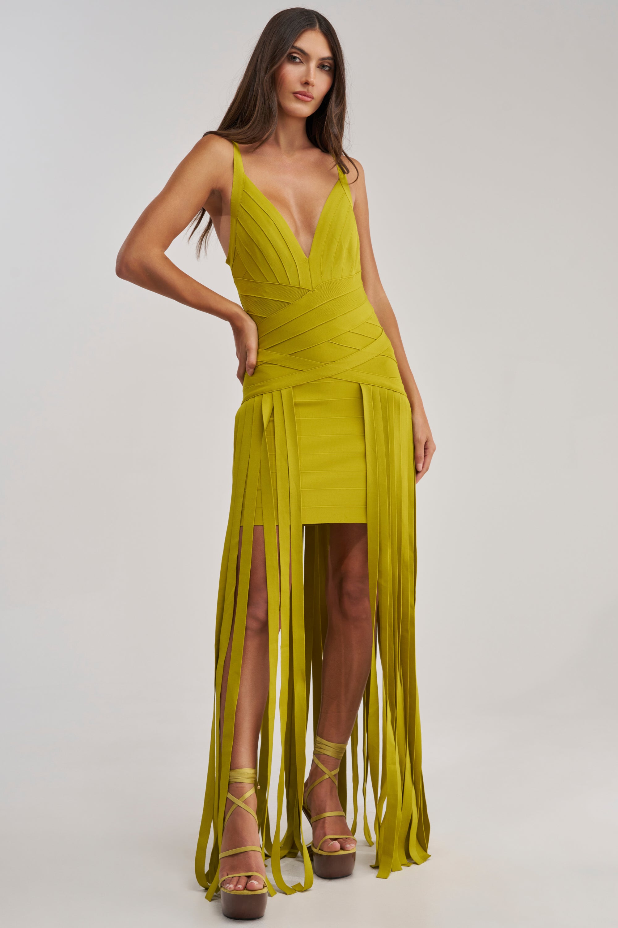 A woman wearing the LUCKY CHARM FRINGE MINI DRESS, a sleeveless chartreuse mini with a deep V-neck, long vertical fringe, and matching platform heels stands against a plain light background.