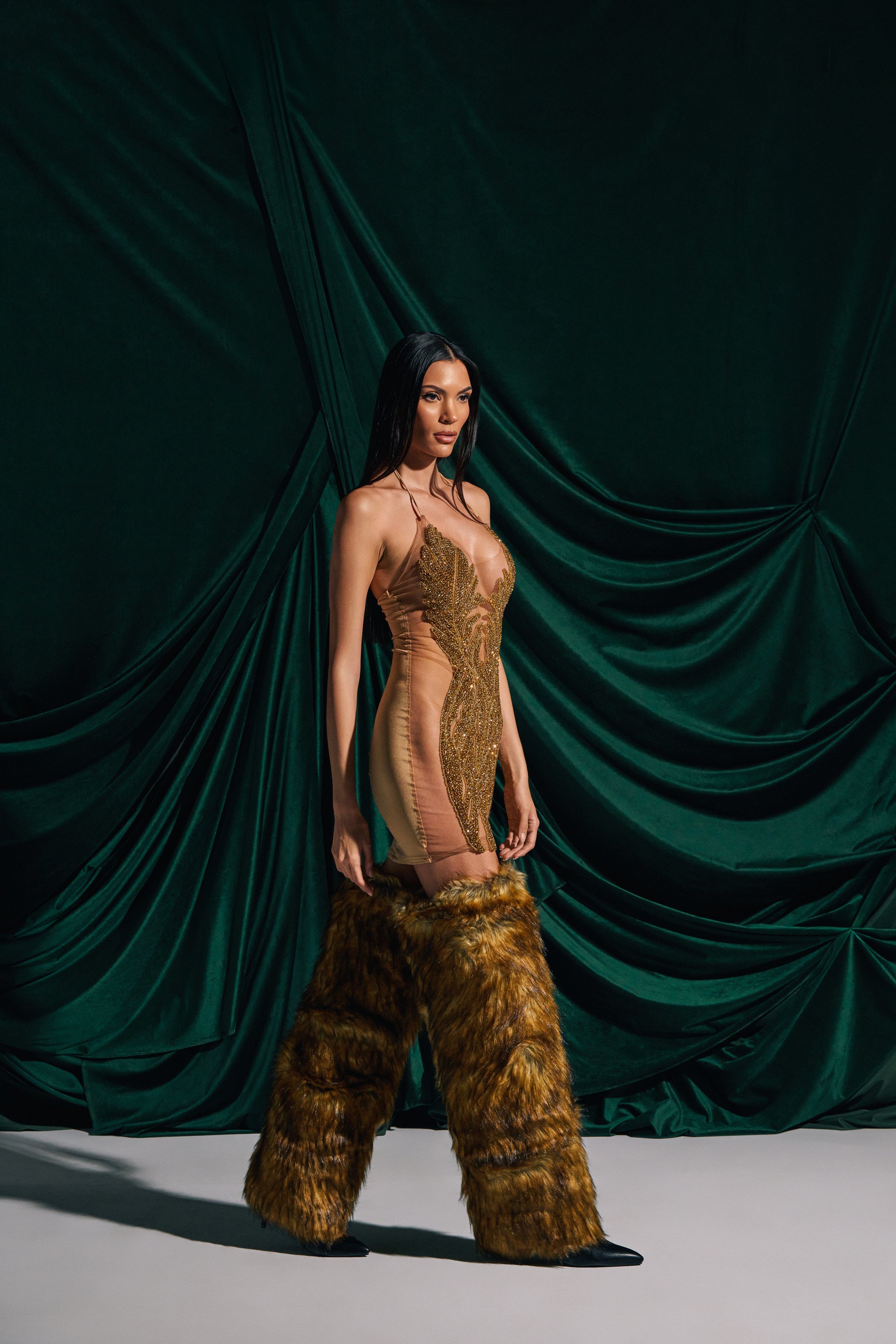 A woman wears the BABY PLEASE MINI DRESS IN GOLD with tall brown furry boots, standing confidently in front of dark green draped fabric and posing with a neutral expression.