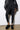 A person clad in a black ensemble, featuring a jacket and ribbed leggings, stands against a white wall. They are wearing the AZALEA WANG DOLORITA BLACK SHERPA CLOG with platform soles. The floor is light brown.