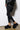 A person embodying the cozy girl look stands against a white wall, dressed in black leggings, a black oversized jacket, and chic AZALEA WANG DOLORITA BLACK SHERPA CLOG slippers. The floor beneath them is a polished brown surface, adding warmth to the stylish ensemble.