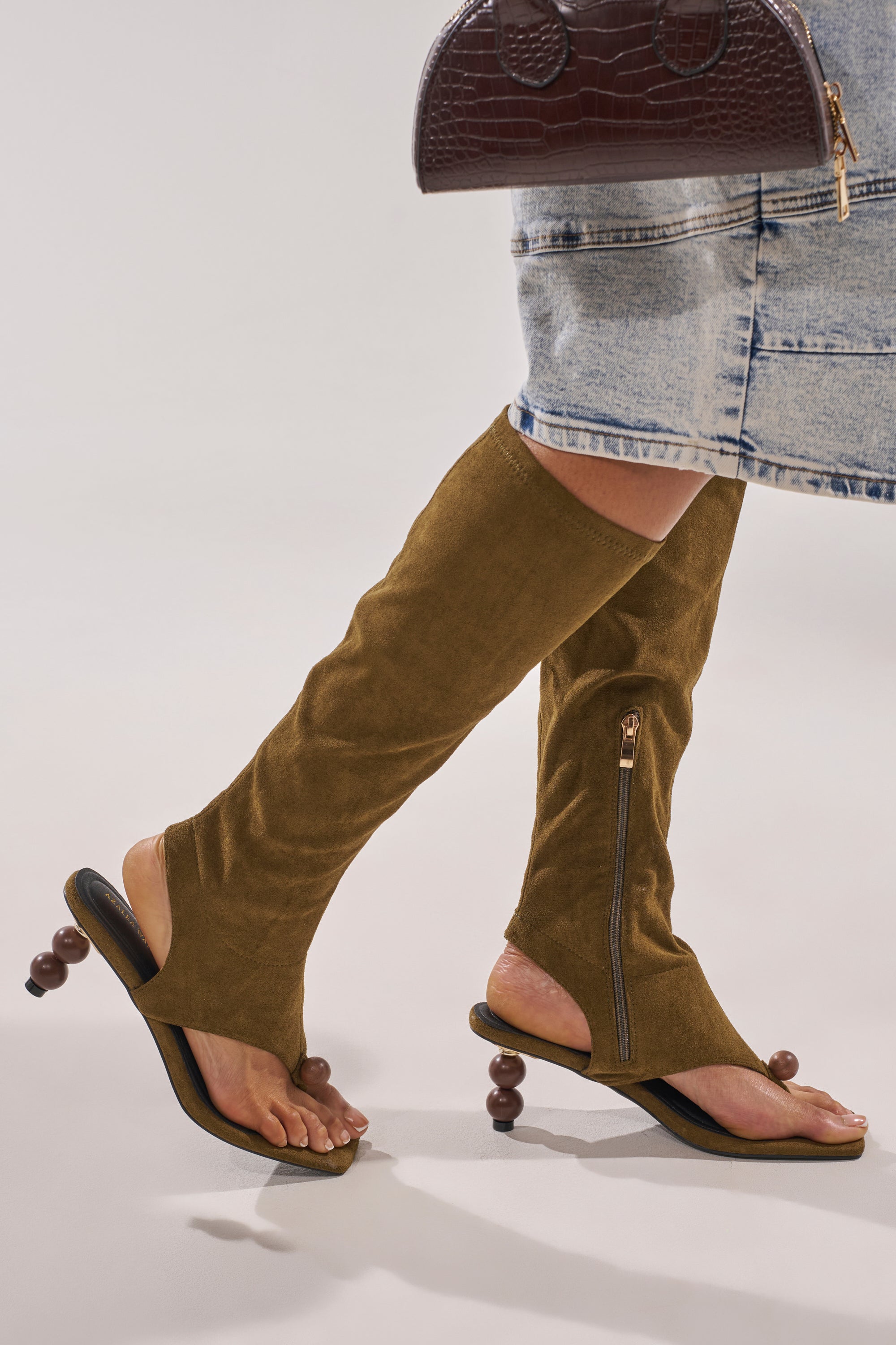 Wearing a denim skirt and the AZALEA WANG DOTTIE Olive Stretch Faux Suede Boot with embellished heel and toe hardware, knee-high shaft, side zippers, and holding a textured brown handbag. 1