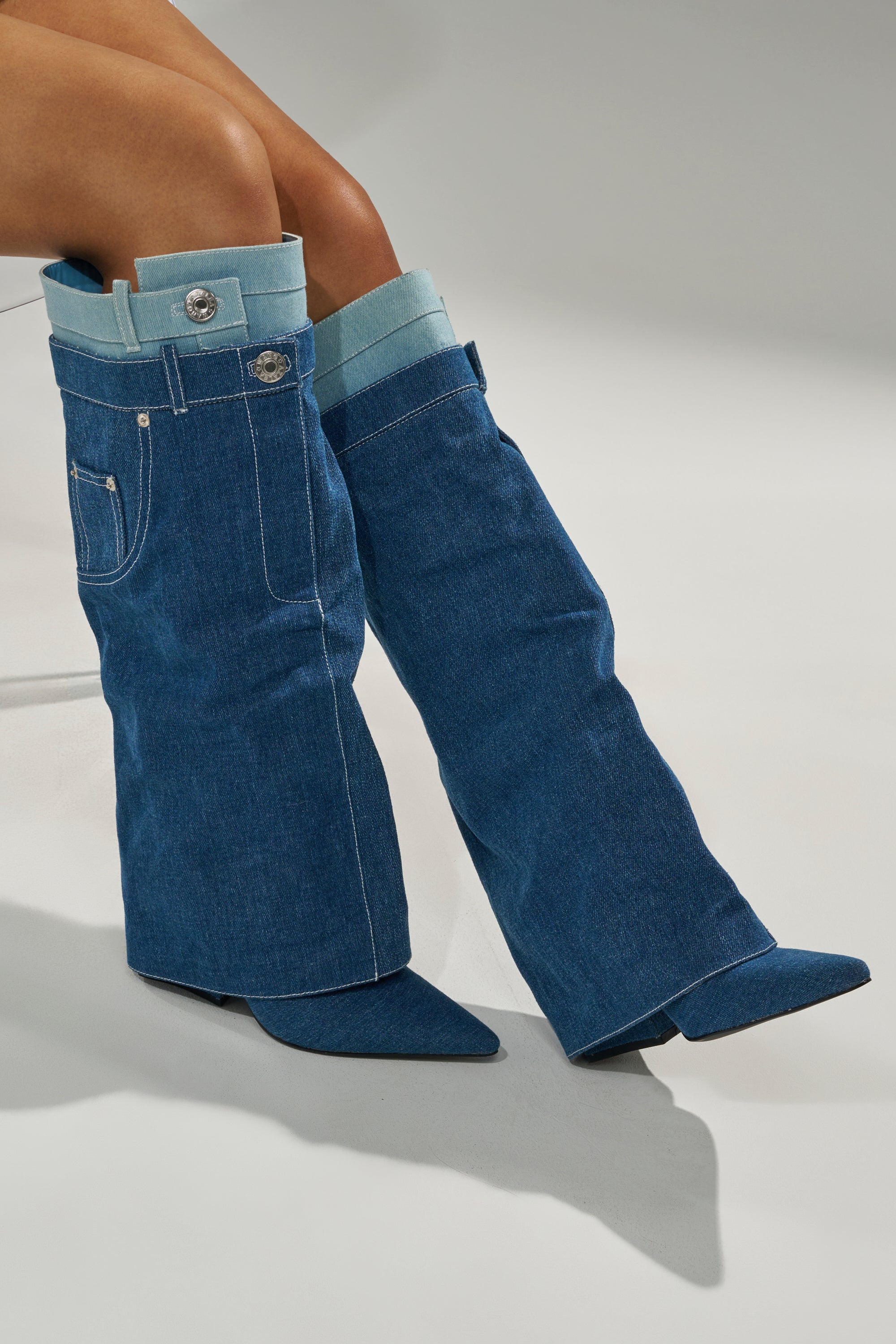 A person wearing the AZALEA WANG DOUBLE FLY DENIM CHUNKY BOOT—knee-high boots styled like jeans with pockets, belt loops, and visible stitching—sits cross-legged against a gray background.
