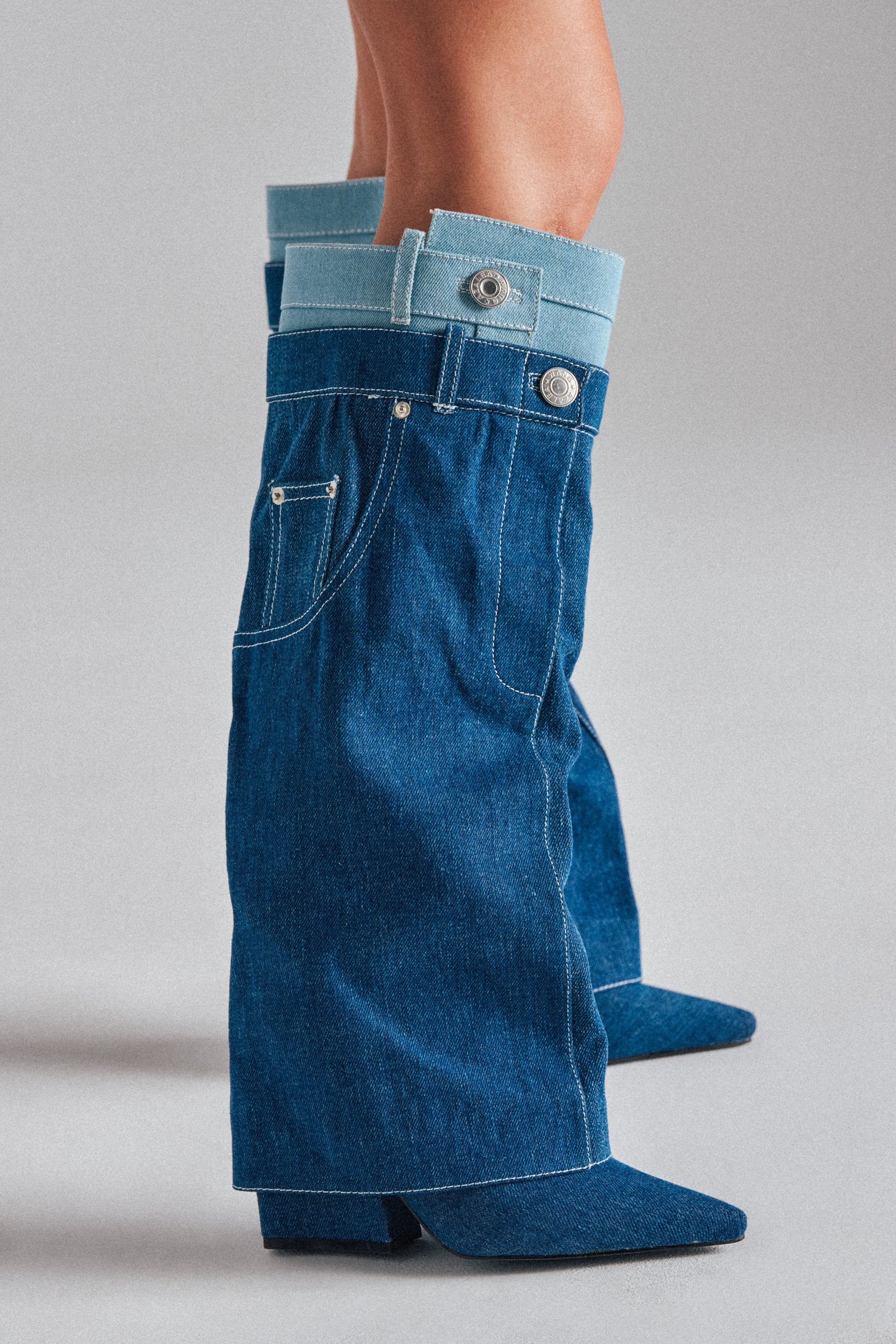 Close-up of lower legs wearing the AZALEA WANG DOUBLE FLY DENIM CHUNKY BOOT—knee-high boots styled like wide-leg jeans with pockets, belt loops, buttons, and a trendy mix of dark and light blue denim. 0