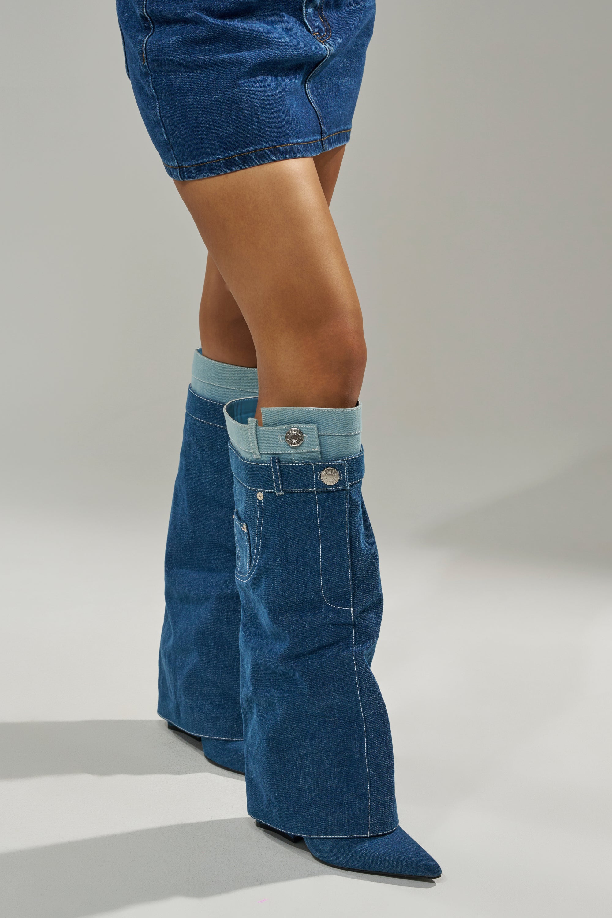 A person wearing a denim mini skirt and AZALEA WANG DOUBLE FLY DENIM CHUNKY BOOTS—knee-high block heel boots styled like upside-down jeans with pockets, belt loops, and buttons—stands on a neutral gray background.