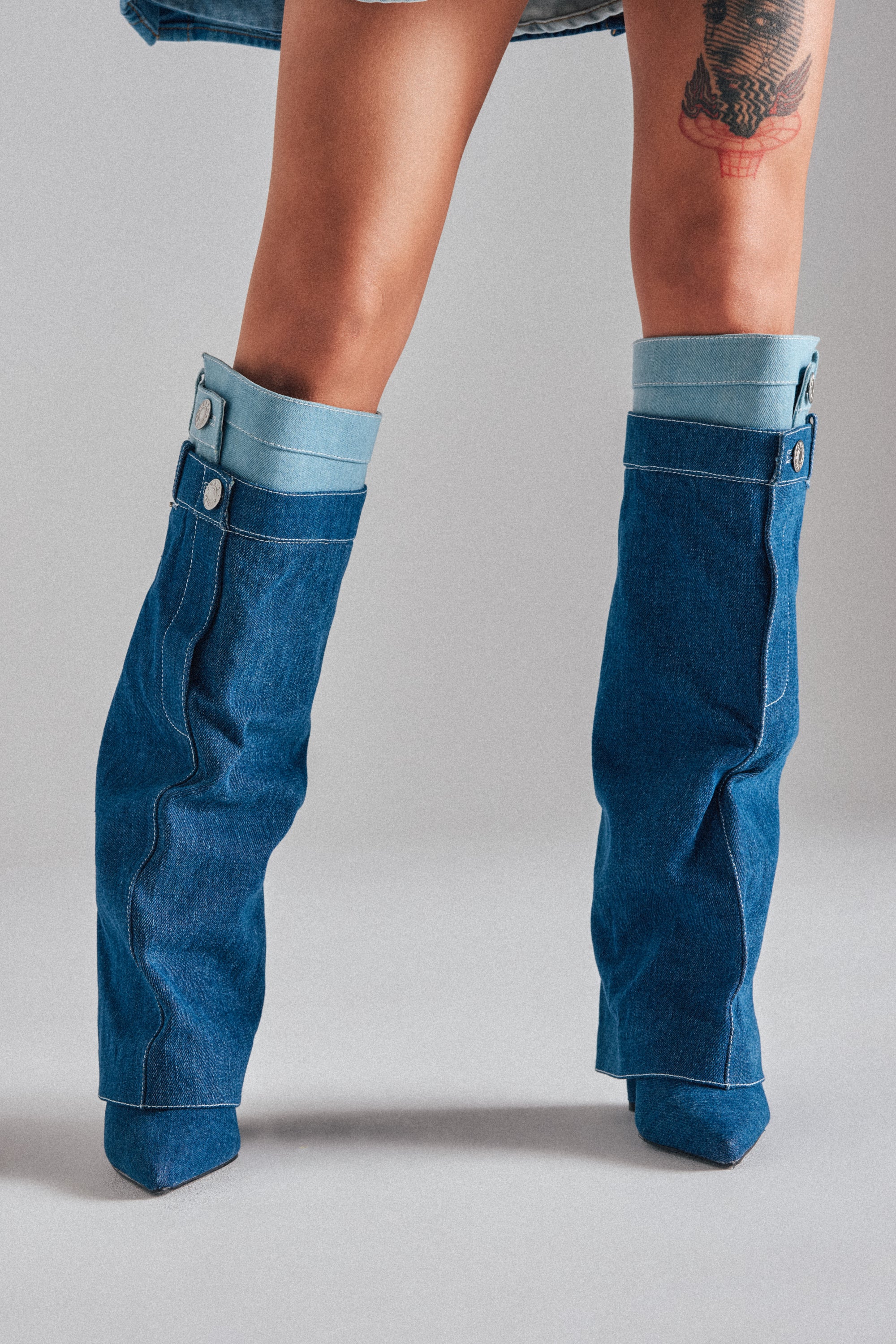 A person with a thigh tattoo wears the AZALEA WANG DOUBLE FLY DENIM CHUNKY BOOT—unique knee-high denim boots with pant leg details, cuffs, and buttons—standing against a plain gray background. 1