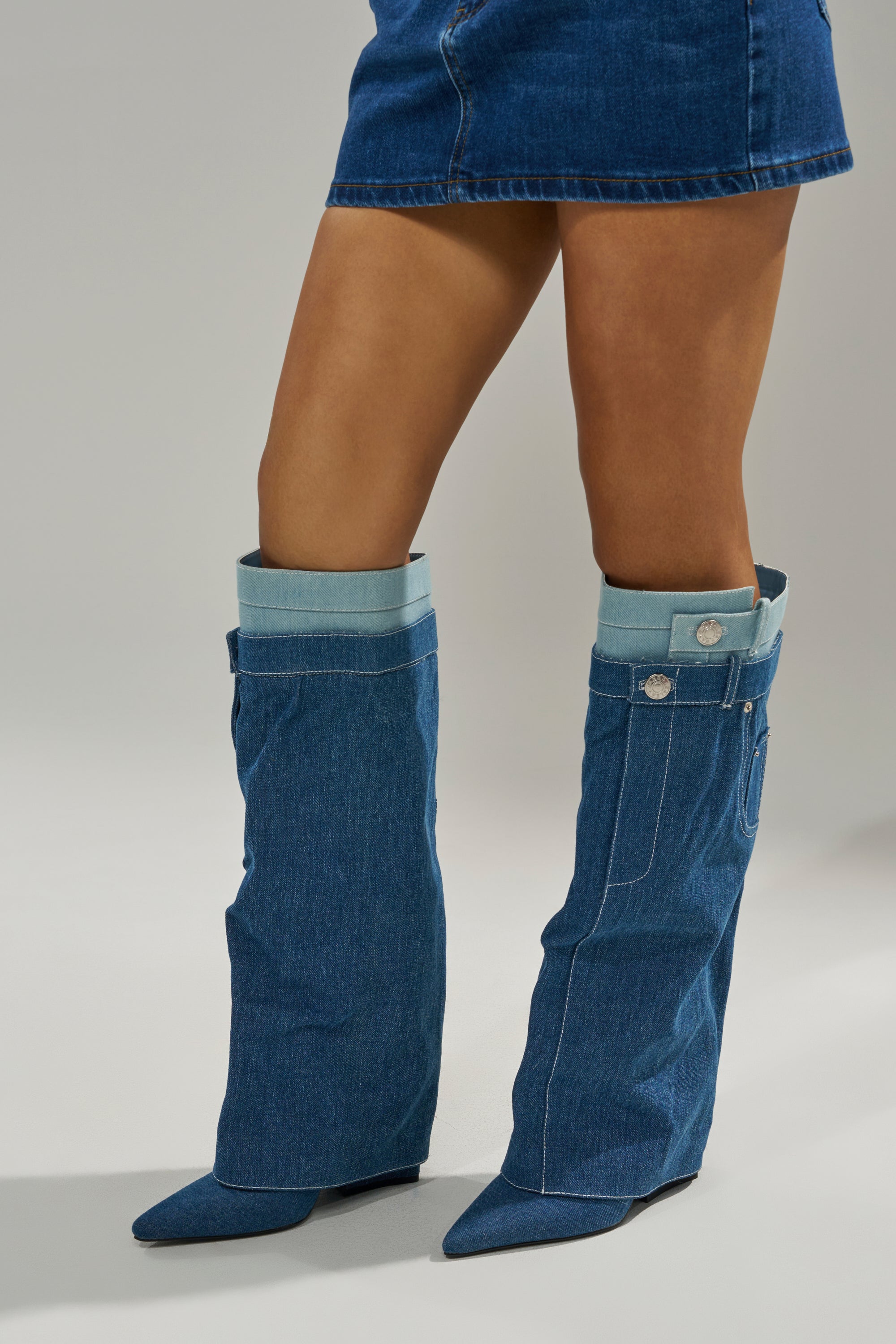 A person wears a short blue denim skirt and AZALEA WANG DOUBLE FLY DENIM CHUNKY BOOT—tall, loose-fitting knee-high boots with pointed toes, silver buttons, and block heels—against a plain light background.