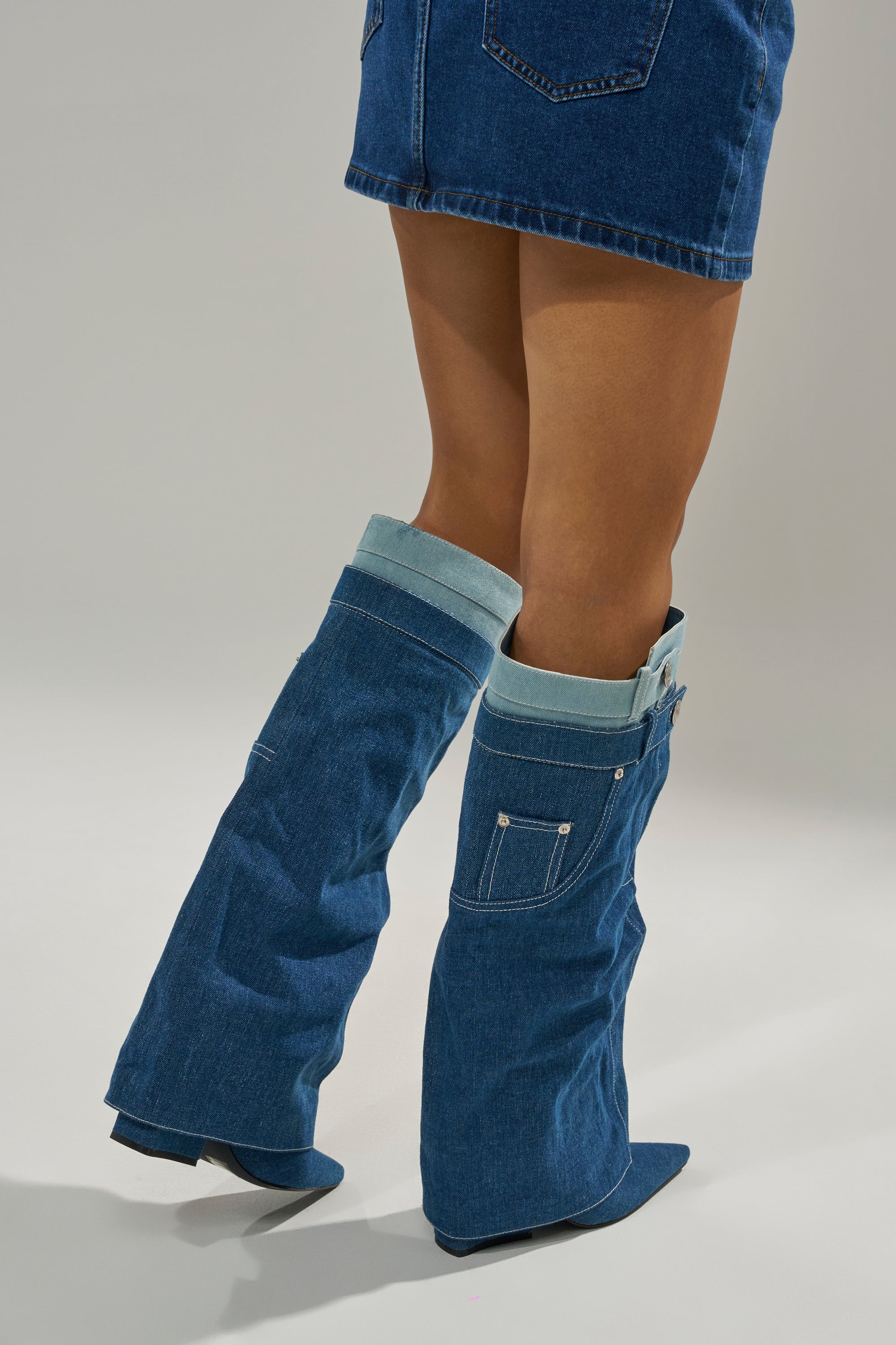 A person wearing a blue denim mini skirt and the AZALEA WANG DOUBLE FLY DENIM CHUNKY BOOT, featuring cuffs and pockets that resemble upside-down jeans, stands on a plain light background.