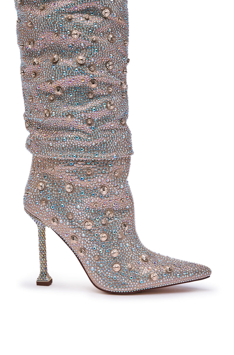 A close-up of the AZALEA WANG DOUGHTY GOLD STILETTO BOOT, featuring sparkling rhinestones, a pointed toe, and a slim gold stiletto heel, set against a crisp white background.