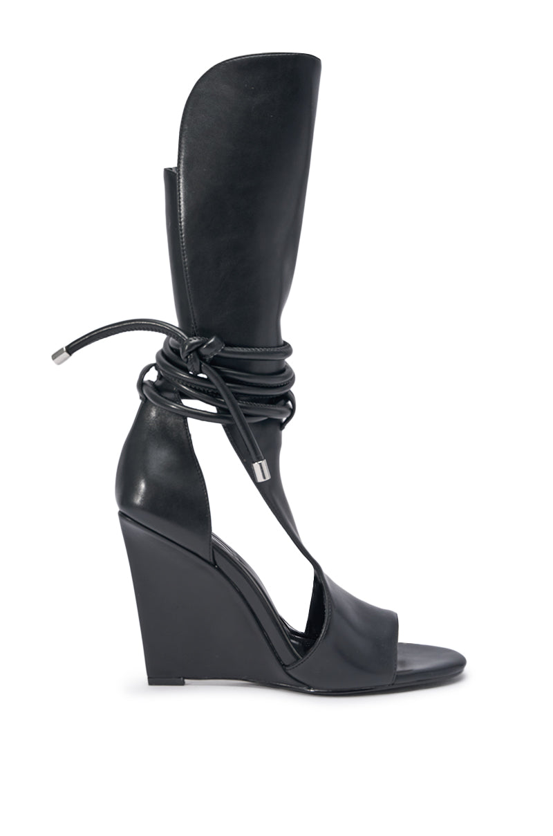 The AZALEA WANG DOUMA BLACK WEDGE LACE UP TALL SHAFT SANDAL features an open toe, high wedge heel, and a tall, structured leather shaft that wraps the ankle with tied leather laces. 2
