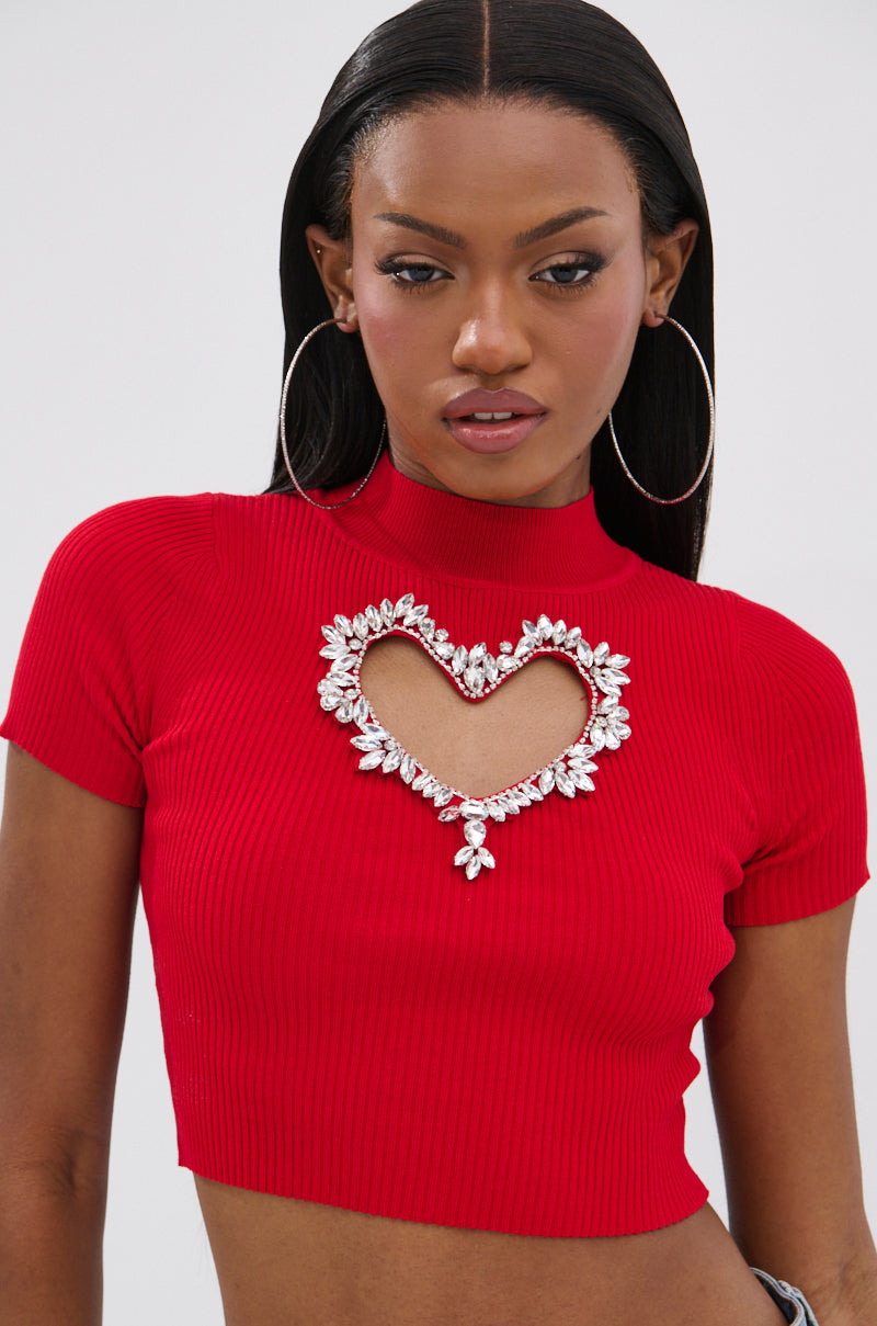 A woman with straight, long dark hair and large hoop earrings wears the HEART YOU TEE IN RED, a fitted short-sleeve red cropped sweater featuring a heart-shaped cutout outlined in rhinestone sparkle.