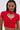 A woman with straight, long dark hair and large hoop earrings wears the HEART YOU TEE IN RED, a fitted short-sleeve red cropped sweater featuring a heart-shaped cutout outlined in rhinestone sparkle.