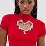 A woman with straight, long dark hair and large hoop earrings wears the HEART YOU TEE IN RED, a fitted short-sleeve red cropped sweater featuring a heart-shaped cutout outlined in rhinestone sparkle.