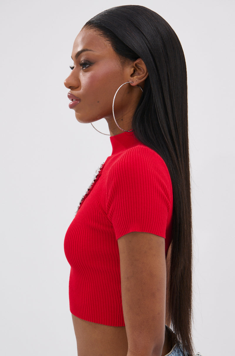 A woman with straight, long dark hair models the HEART YOU TEE IN RED—a fitted, short-sleeve cropped sweater with a heart-shaped cut-out—paired with large hoop earrings, shown in profile against a plain light background.