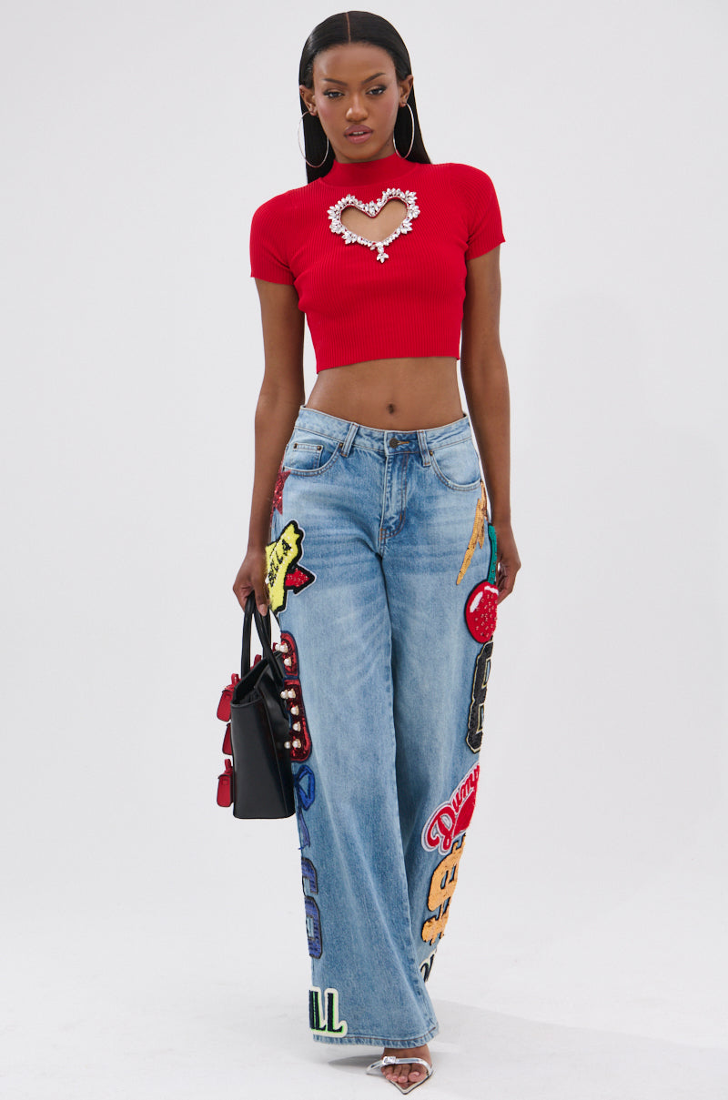 A woman wears the HEART YOU TEE IN RED with a heart cut-out, paired with light blue wide-leg jeans with rhinestone patches, a small black handbag, hoop earrings, and heeled sandals.