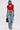 A woman wears the HEART YOU TEE IN RED with a heart cut-out, paired with light blue wide-leg jeans with rhinestone patches, a small black handbag, hoop earrings, and heeled sandals.