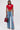 A woman wears the HEART YOU TEE IN RED with rhinestones, heart cutout, and pearl trim, paired with wide-leg blue jeans featuring colorful patches, clear heels, large hoop earrings, and a black handbag adorned with red purses.