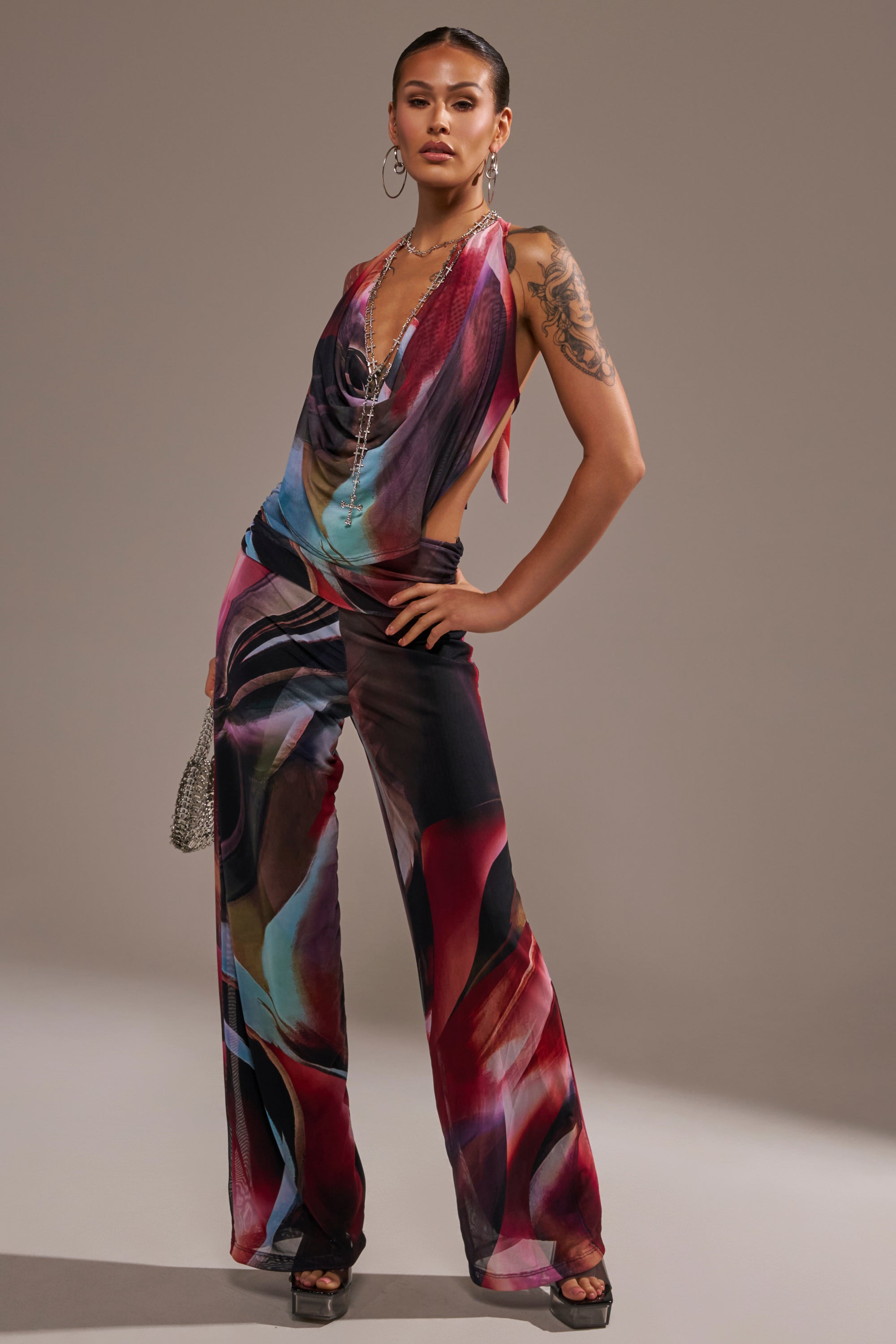 A woman exudes confidence in the MAIN CHARACTER ENERGY COWL NECK JUMPSUIT with wide-leg pants, accessorized with hoop earrings, layered necklaces, a chic handbag, and open-toe heels; her hair is slicked back for a stylish finish. 0