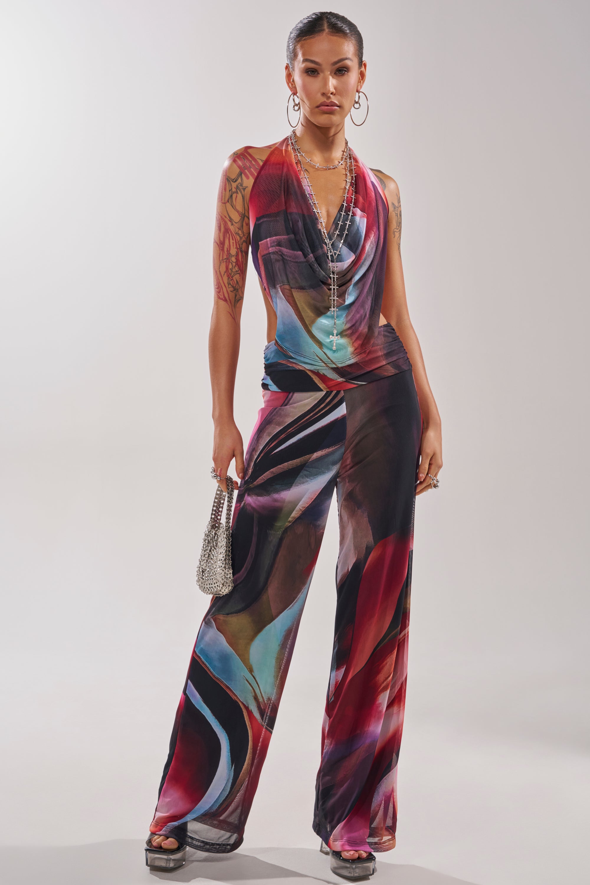 A woman stands confidently in the MAIN CHARACTER ENERGY COWL NECK JUMPSUIT, a vibrant abstract print halter design with flared pants. She accessorizes with a sparkling silver handbag, layered necklaces, hoop earrings, and sandals. 1